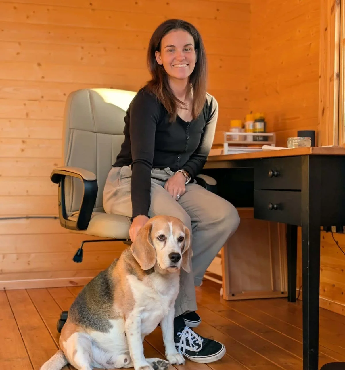 A woman with brown hair sitting on a chair in a wooden room, smiling, with her hand on a Beagle dog sitting on the floor in front of her.