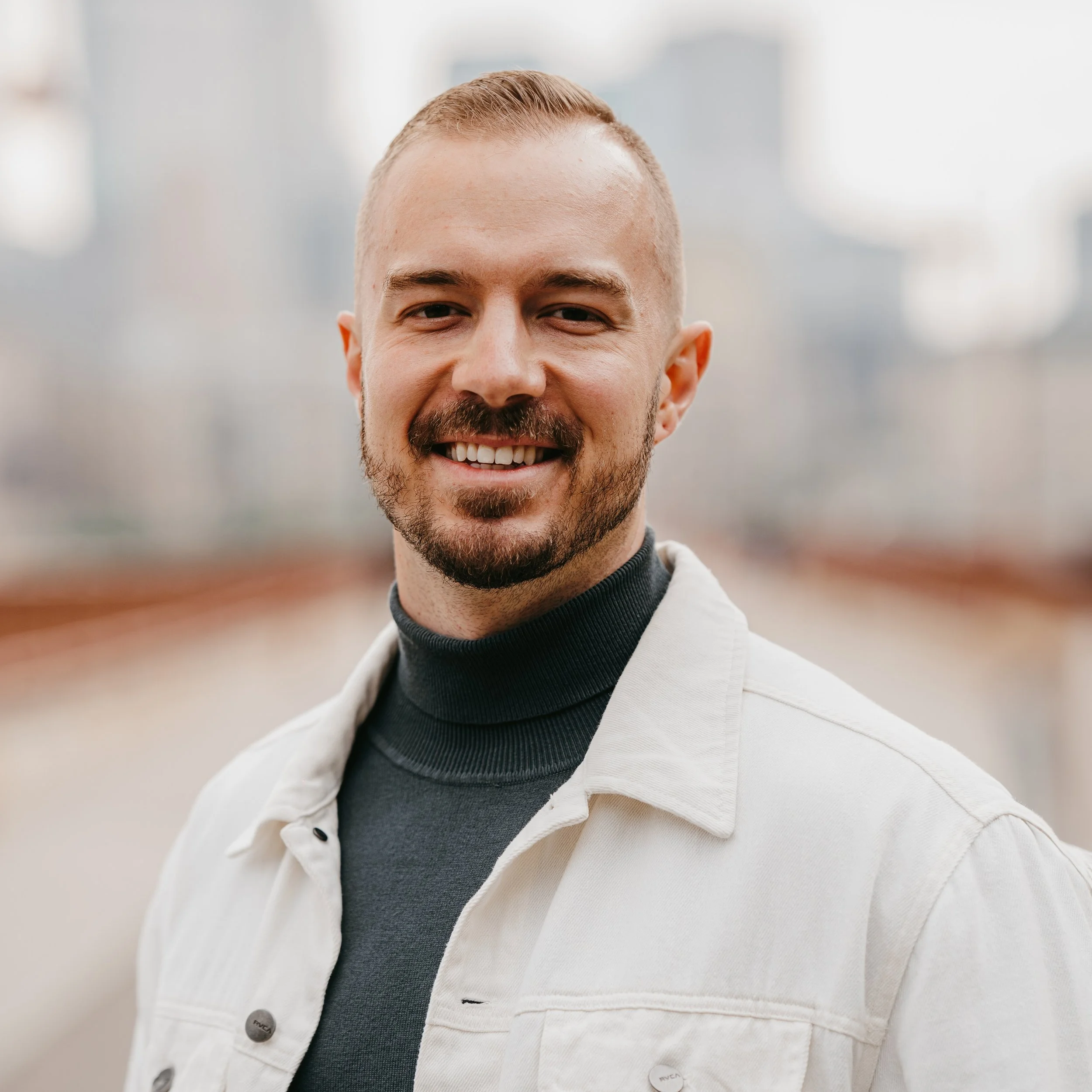 Close-up of a young man with a short beard and smiling, wearing a black turtleneck and a white jacket, outdoors with blurred background.