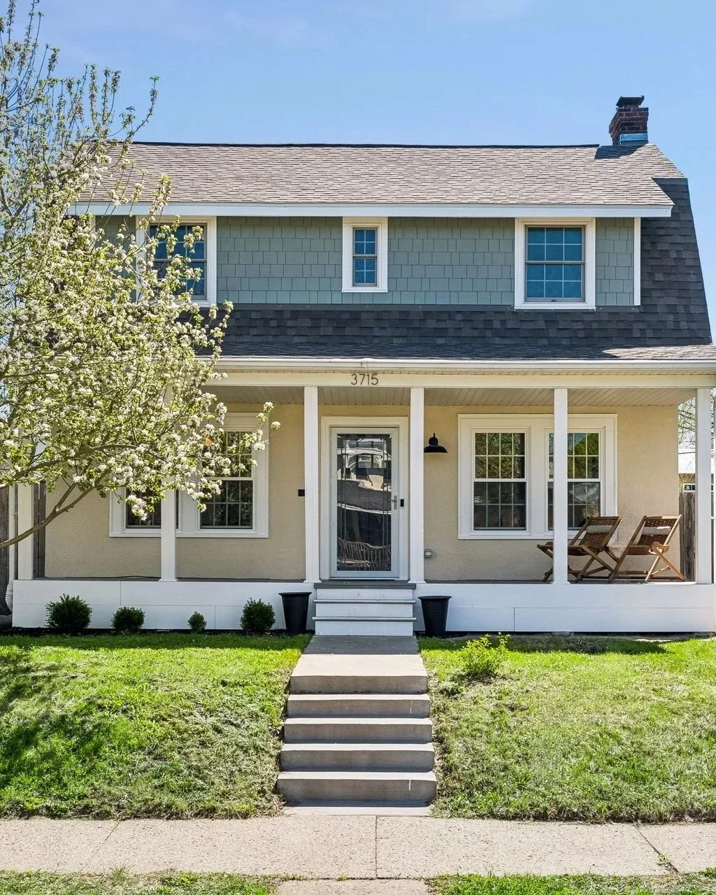 Front view of a two-story house with a porch, cream-colored walls, gray roof, and four windows, with a small tree on the left and a grassy lawn in front.