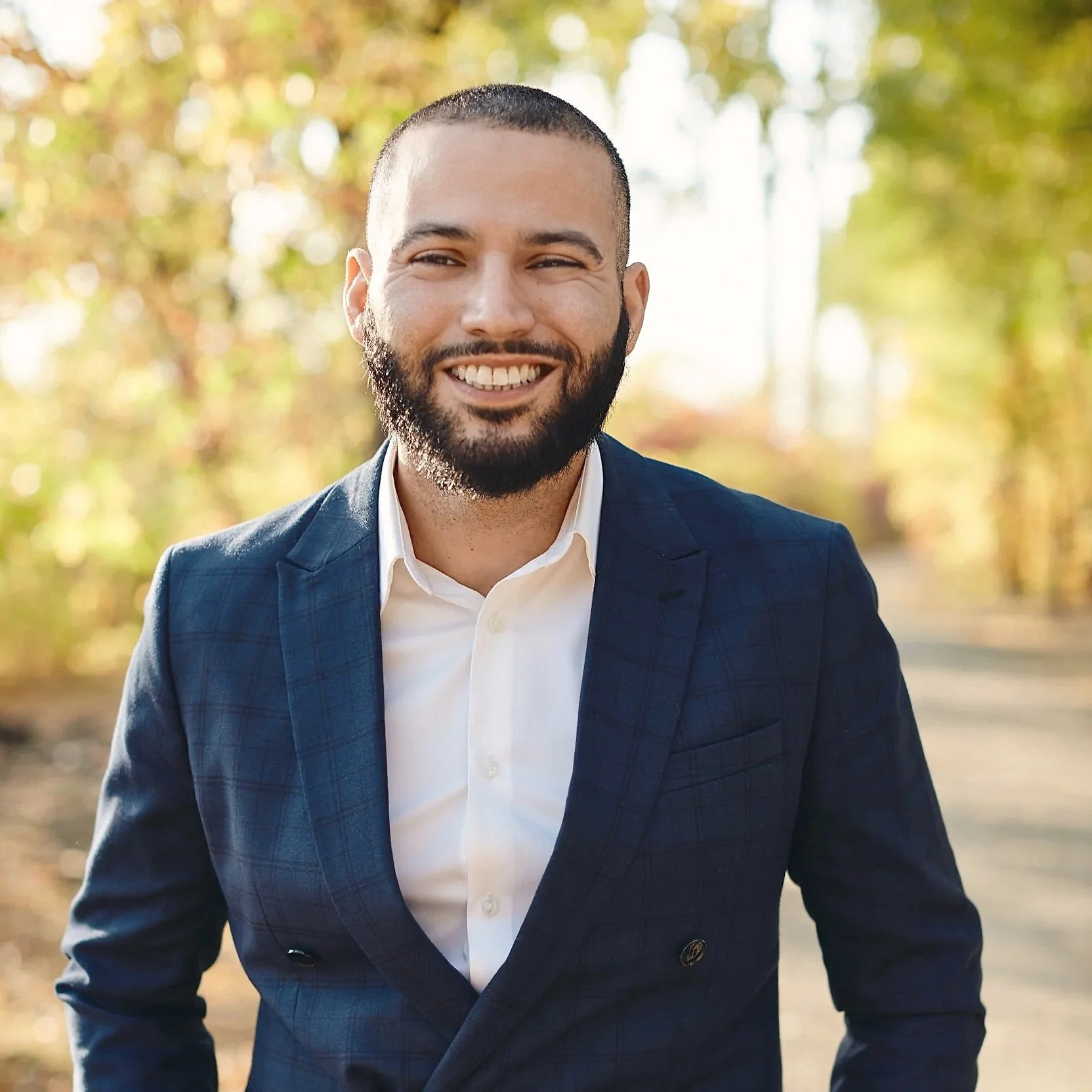 A smiling man in a navy blue suit and white shirt outdoors with blurry trees in the background.