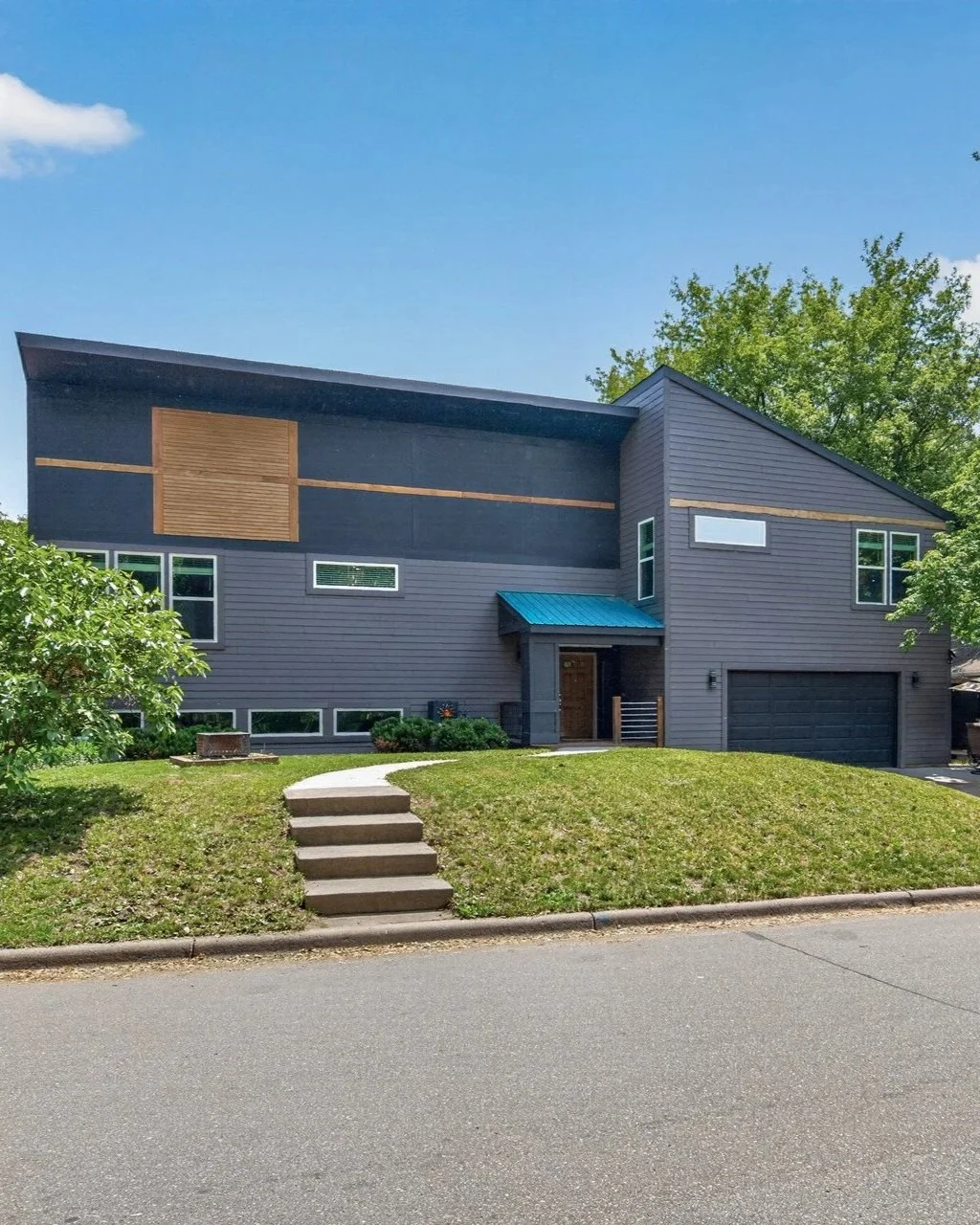 Modern two-story house with gray siding, a small front porch with a blue metal roof, and a front yard with steps leading up from the sidewalk. The house has multiple windows and is surrounded by green trees and grass.