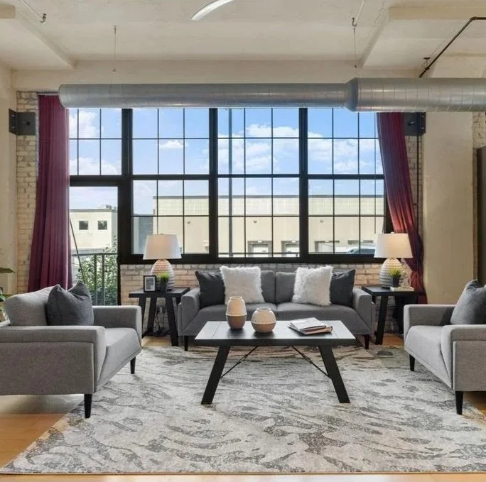Modern living room with large window, gray sofas, side tables with lamps, decorative pillows, a coffee table with vases, and a patterned rug.