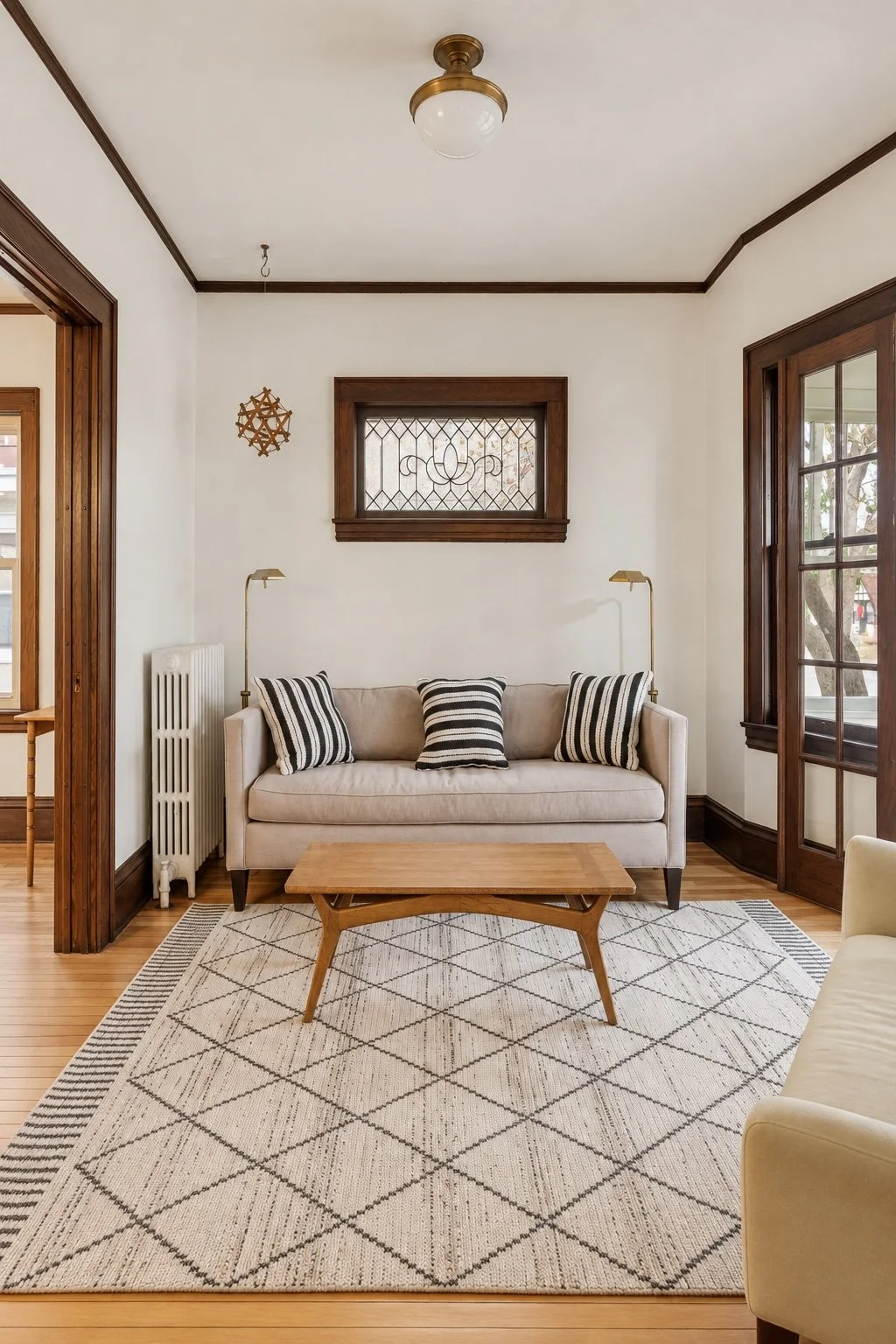 Living room with a beige sofa with black and white striped pillows, a wooden coffee table, a white area rug with a diamond pattern, dark wood window and door frames, a small window with decorative glass, and a wall-mounted gold lamp.