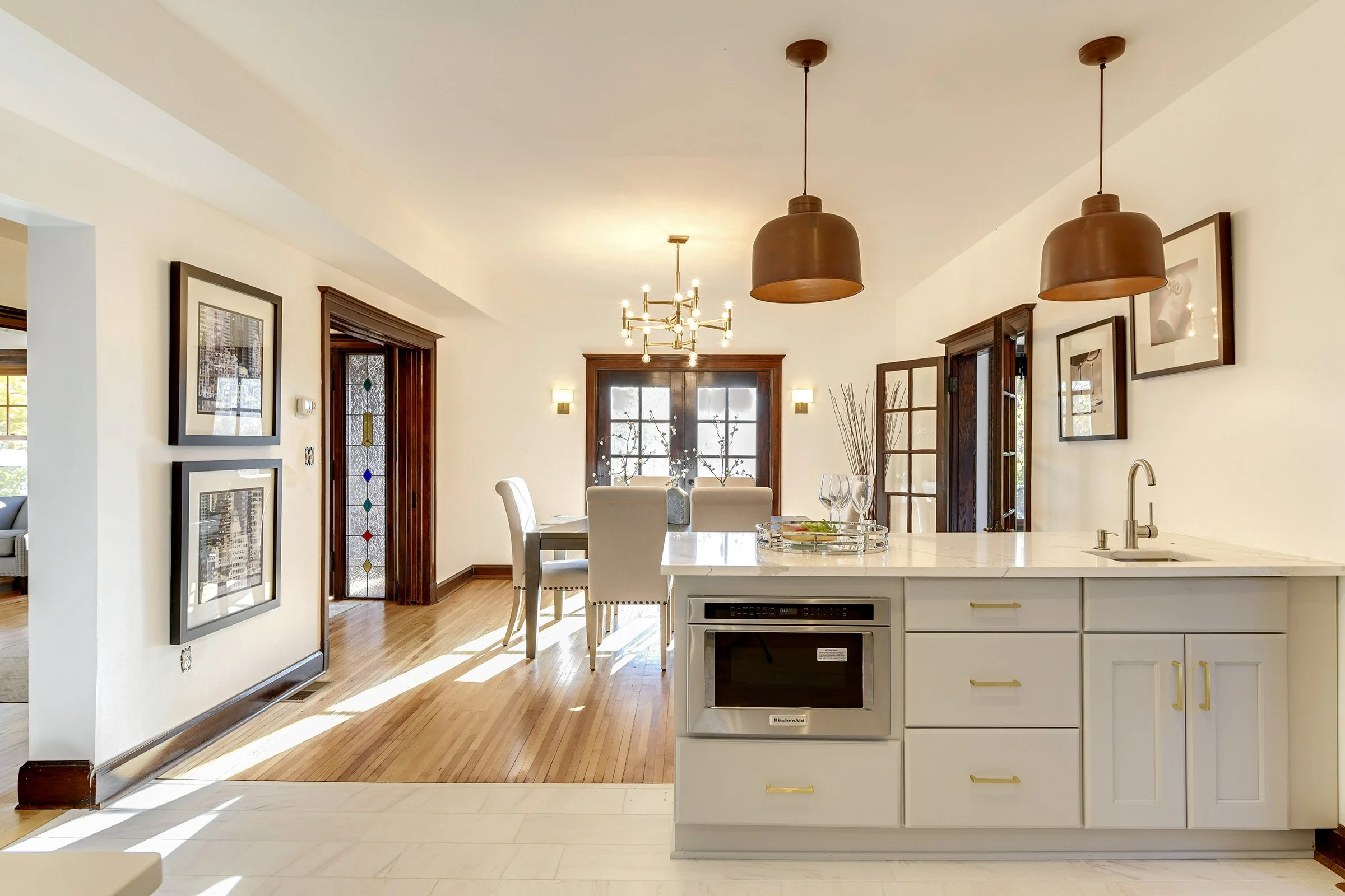 A bright kitchen and dining area with white cabinetry, a white marble island, and hardwood floors. The space has modern hanging brown pendant lights and a chandelier over the dining table. There are framed artworks on the wall, and natural light streams through windows and glass-paneled double doors.