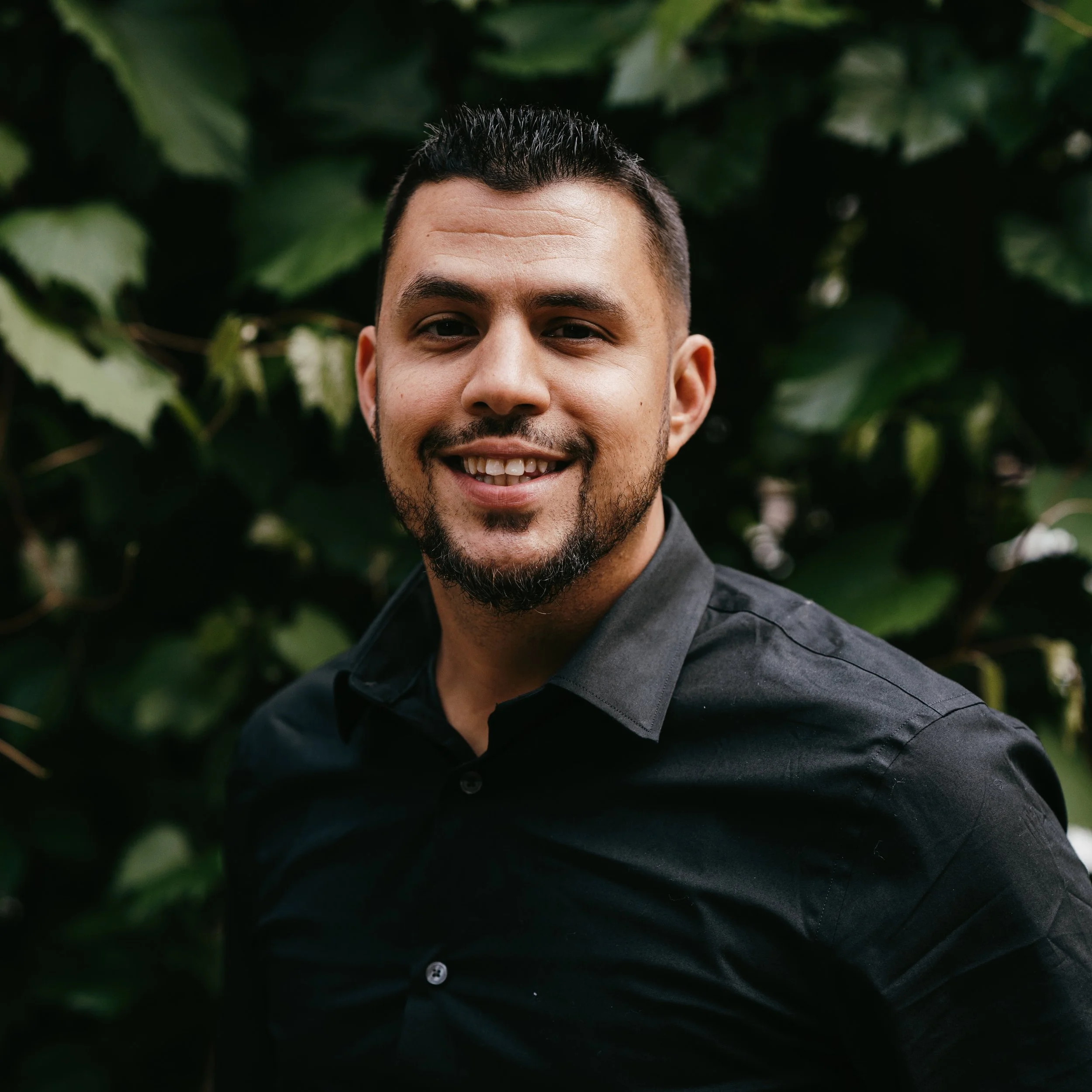 A smiling man with dark hair, a beard, and light brown skin, wearing a black shirt, standing outdoors with green foliage in the background.