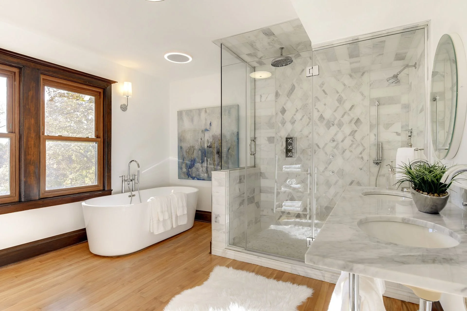 A modern bathroom with a white bathtub, wooden window frame, a glass shower enclosure with marble tiles, a round mirror, a white sink, and a potted plant, illuminated by natural light and modern ceiling lighting.