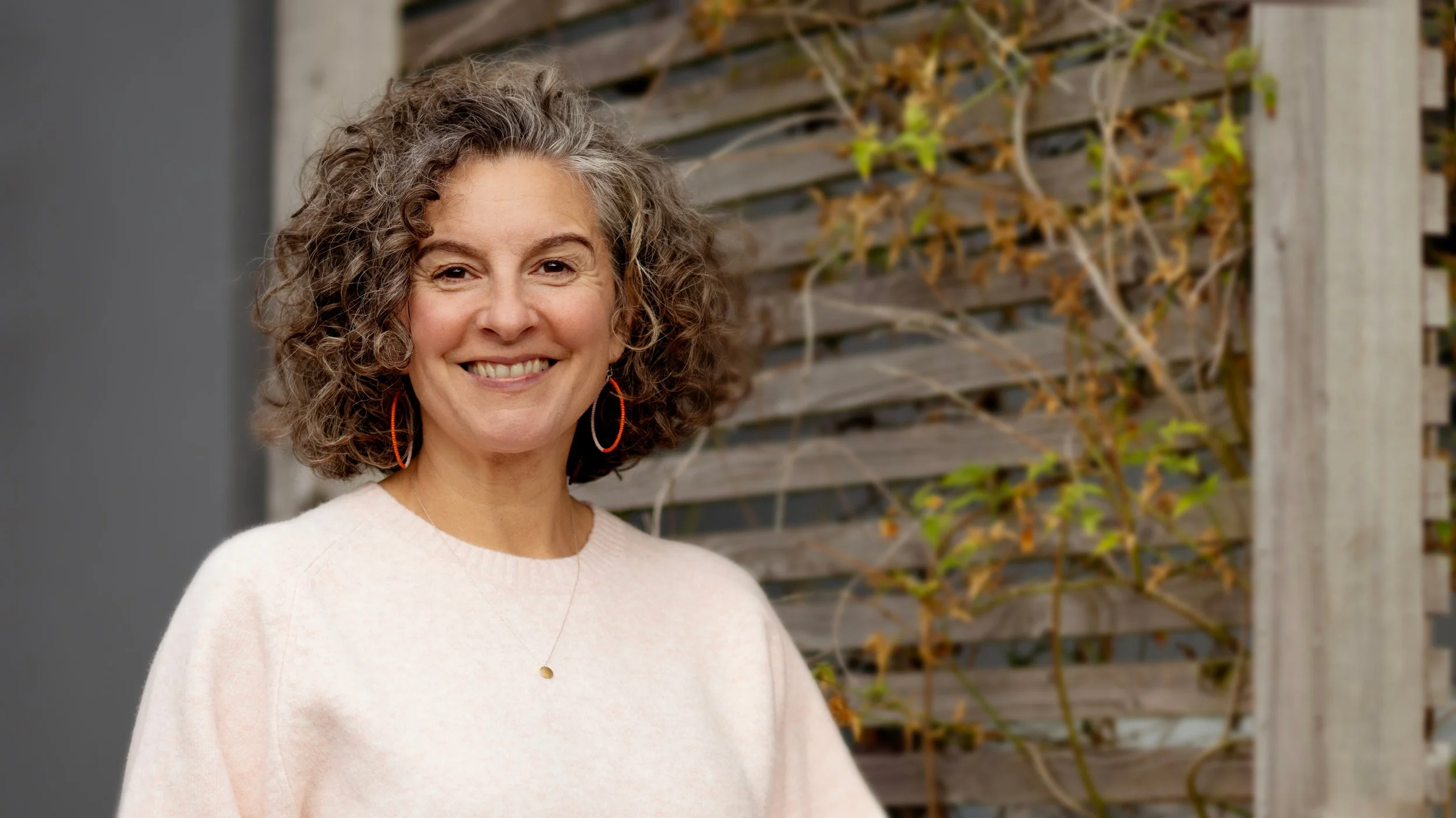 Photo of Helen Drane hypnotherapist, standing in front of a fence, wearing a light pink jumper and smiling