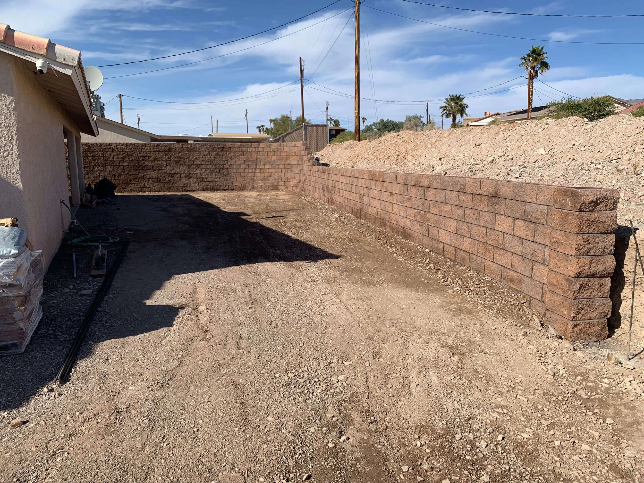 A backyard under construction with a dirt ground, a brick wall, and a house with a satellite dish and security camera. Utility poles and wires are visible, with palm trees and neighboring houses in the background under a partly cloudy sky.