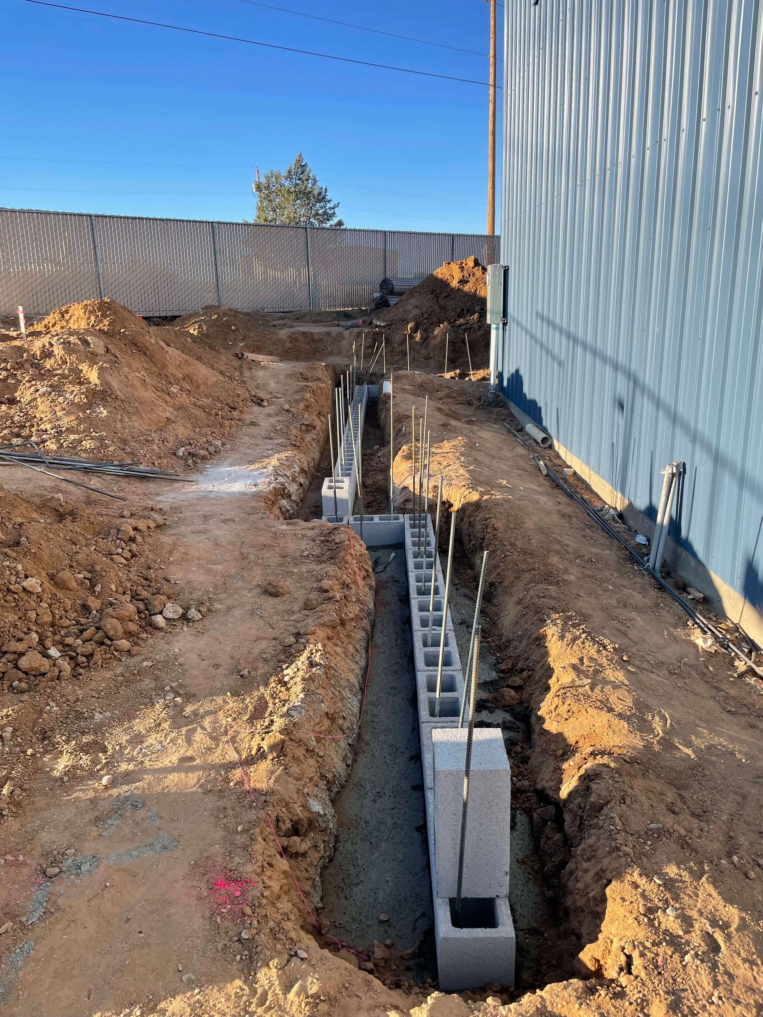 Construction site with dug trench, concrete blocks, and rebar, next to a metal building, under a clear blue sky.