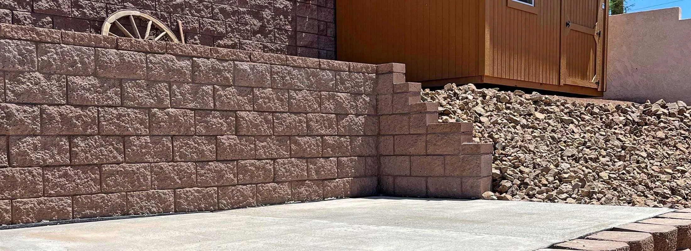A brick retaining wall with a decorative wagon wheel embedded near the top, a gravel slope, a wooden shed, and a concrete sidewalk in the yard.