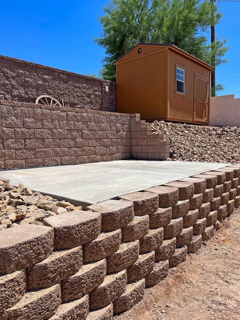 Backyard with terraced brick retaining wall, concrete slab patio, small wooden shed, and clear blue sky.