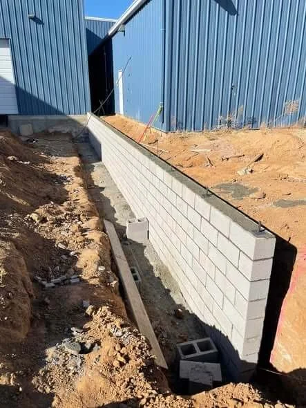 Construction site with a partially built cinder block wall next to a blue metal building, with dirt and construction materials on the ground.