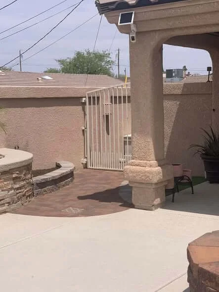 Residential backyard patio with a stucco wall, a fenced gate, and a covered porch area with a potted plant.