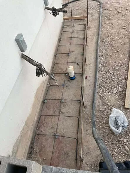 Concrete pathway with rebar and wire laying along a building wall, with electrical cords, a bottle, and construction debris nearby.