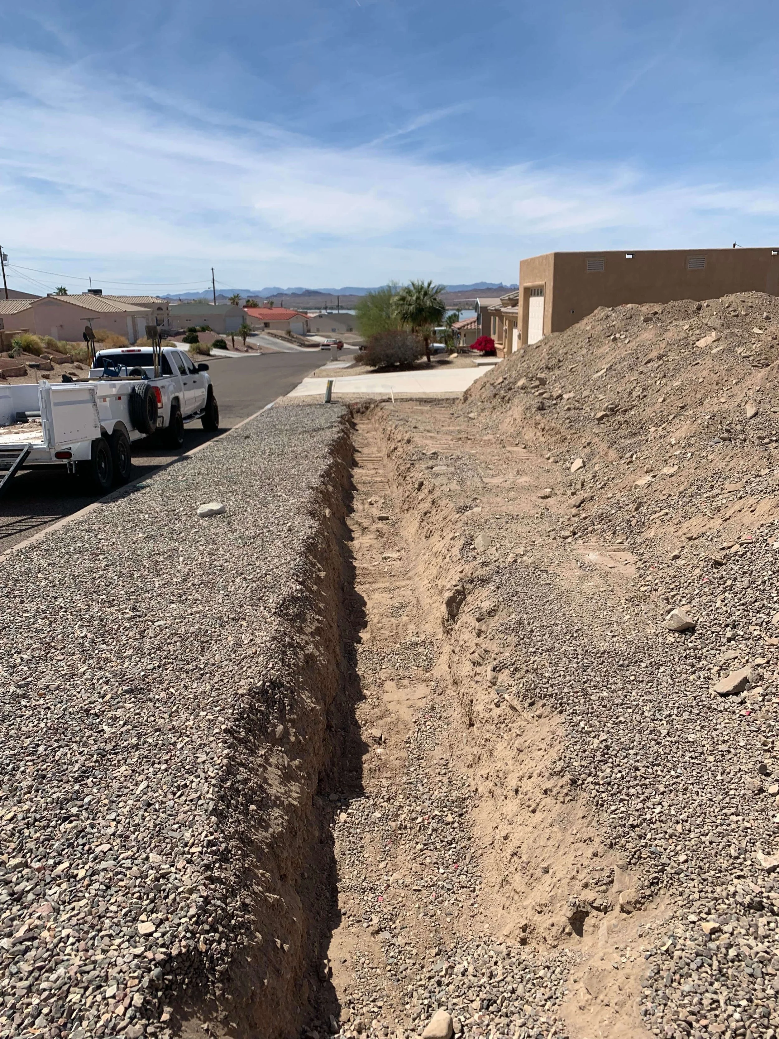 A sidewalk construction site with a narrow trench in the ground, gravel piled on one side, and residential houses in the background under a blue sky.