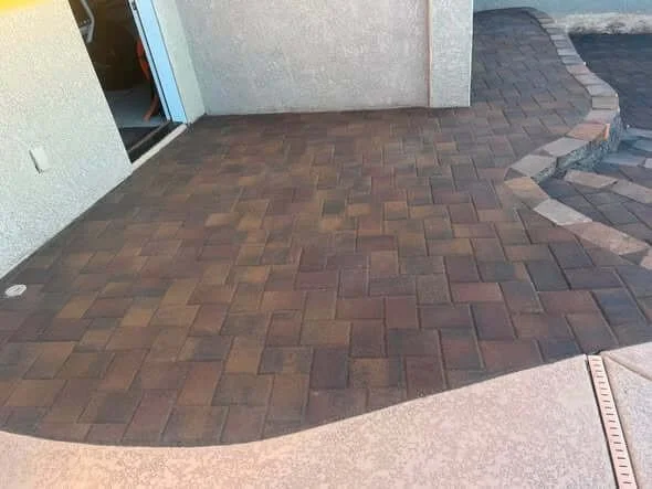 Patterned brick pavers form a walkway outside a building, with a concrete curb and a glass door in the background.