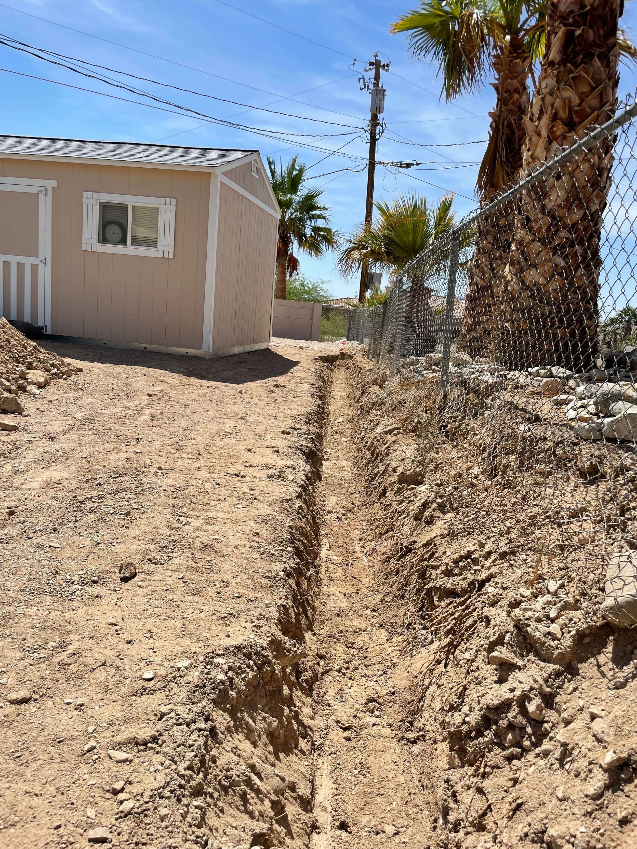 A dirt path with a deep trench dug along the right side, lined with rocks and dirt, in a yard with a beige house, palm trees, a chain-link fence, and power lines against a blue sky.