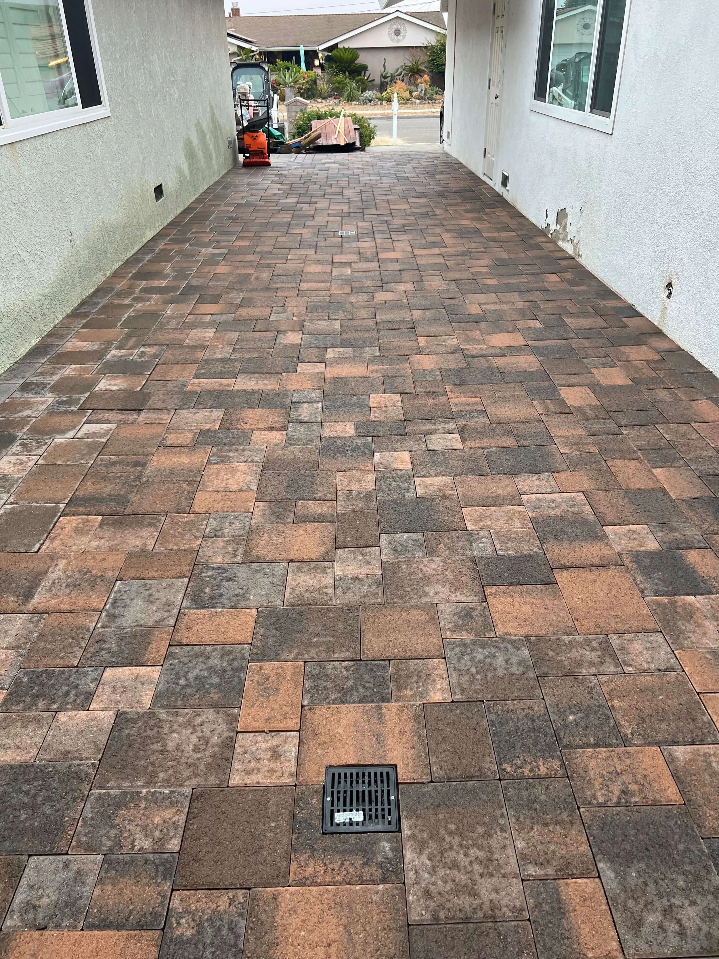 A paved outdoor area with interlocking bricks in shades of brown and gray, a drain cover at the center, and part of a house wall with windows and some peeling paint or damage, with a view of a garden and street beyond.