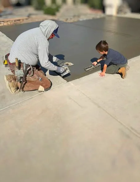 A construction worker and a young boy are both kneeling on a roof, installing or repairing a flat roof surface.