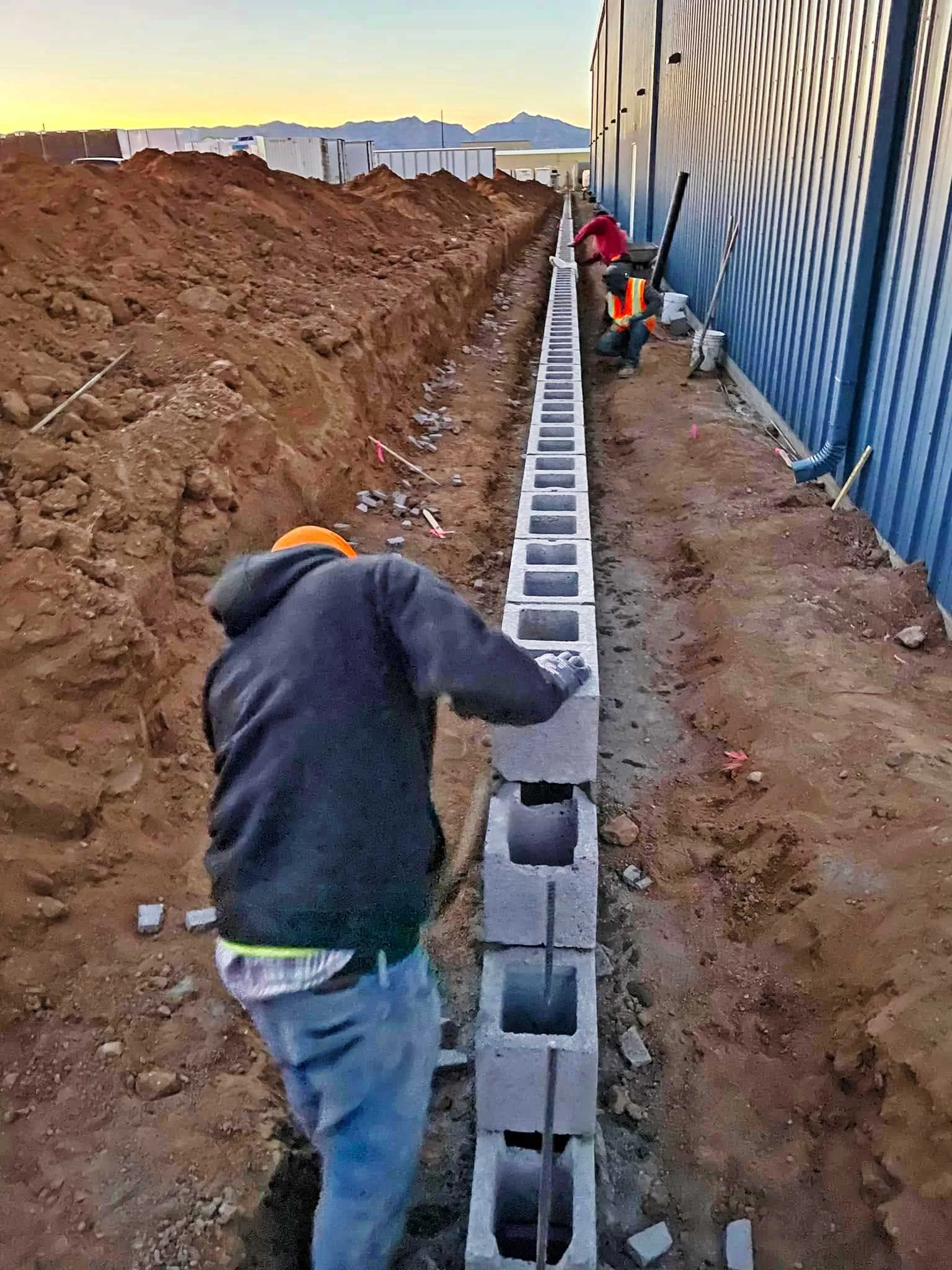 Workers laying cinder blocks along a construction site with dirt piles and a blue metal building wall, mountains in the background at sunset.