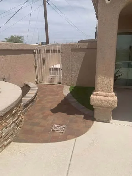 A small outdoor area with a curved brick pathway, a gate, and part of a house with an archway and a glass door, under a cloudy sky.
