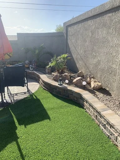 Backyard with artificial grass, a curved brick retaining wall, small palm trees, decorative rocks, and outdoor lighting fixtures.