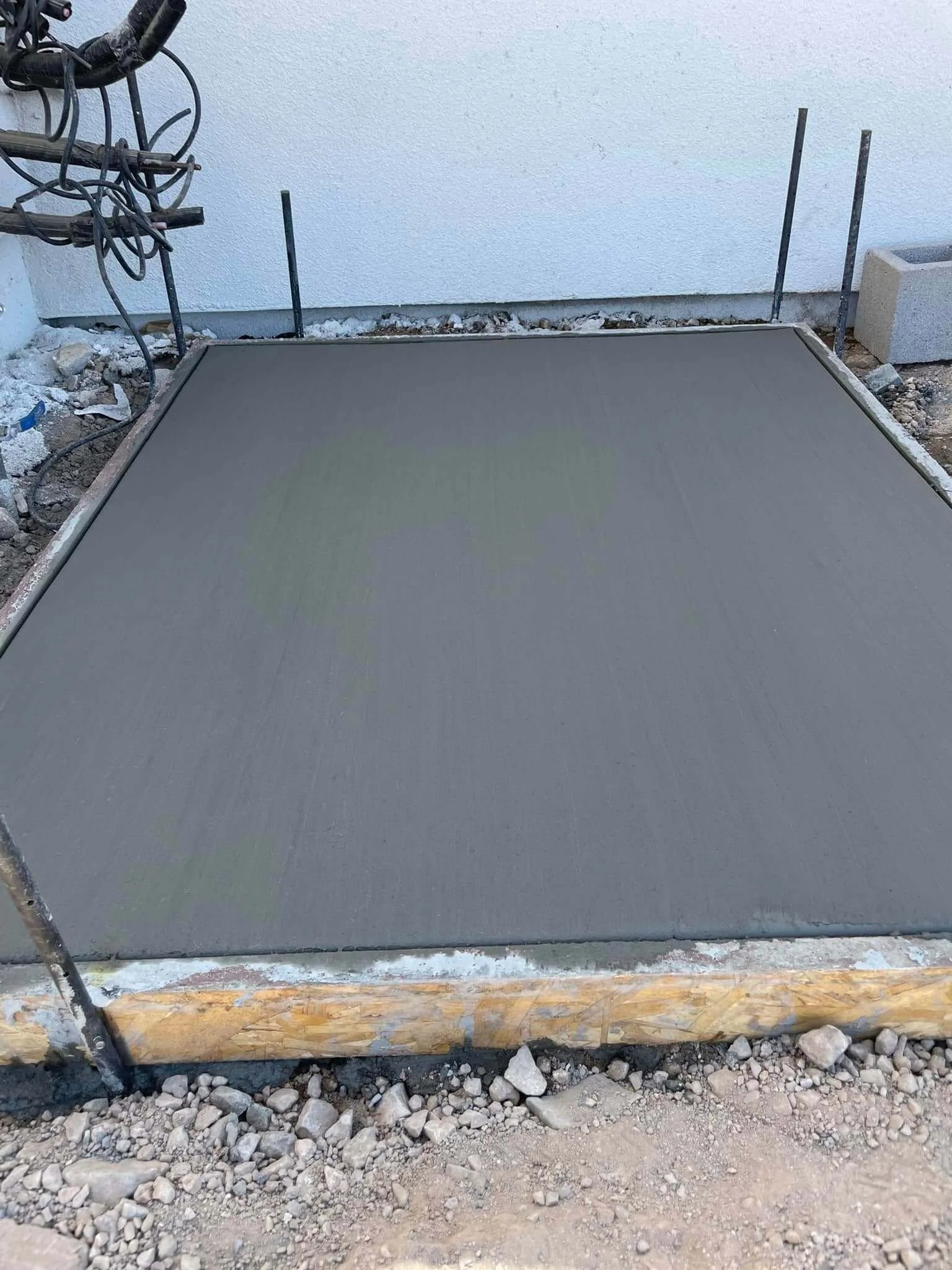 Freshly poured concrete slab on a construction site, with gravel and dirt surrounding it, and wooden formwork at the edges.