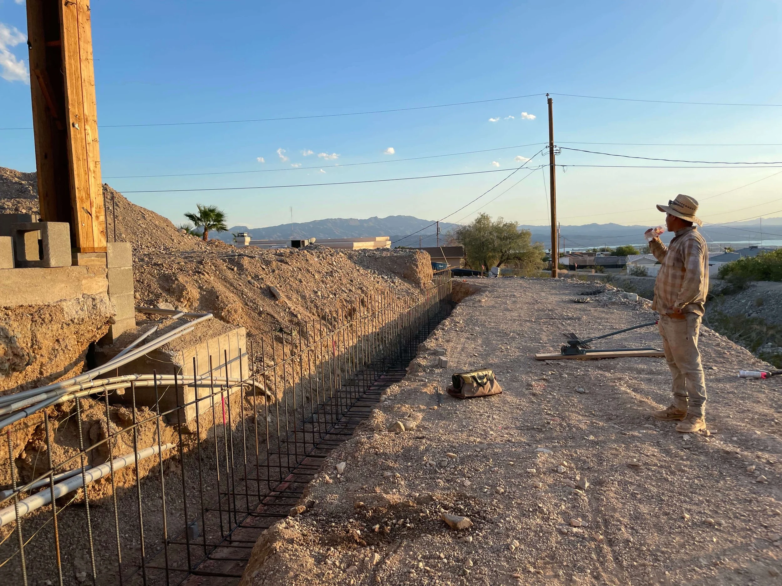 A construction worker wearing a straw hat, plaid shirt, and mask standing by a rebar foundation with electrical conduit pipes, in a rural desert area with mountains in the background during late afternoon.