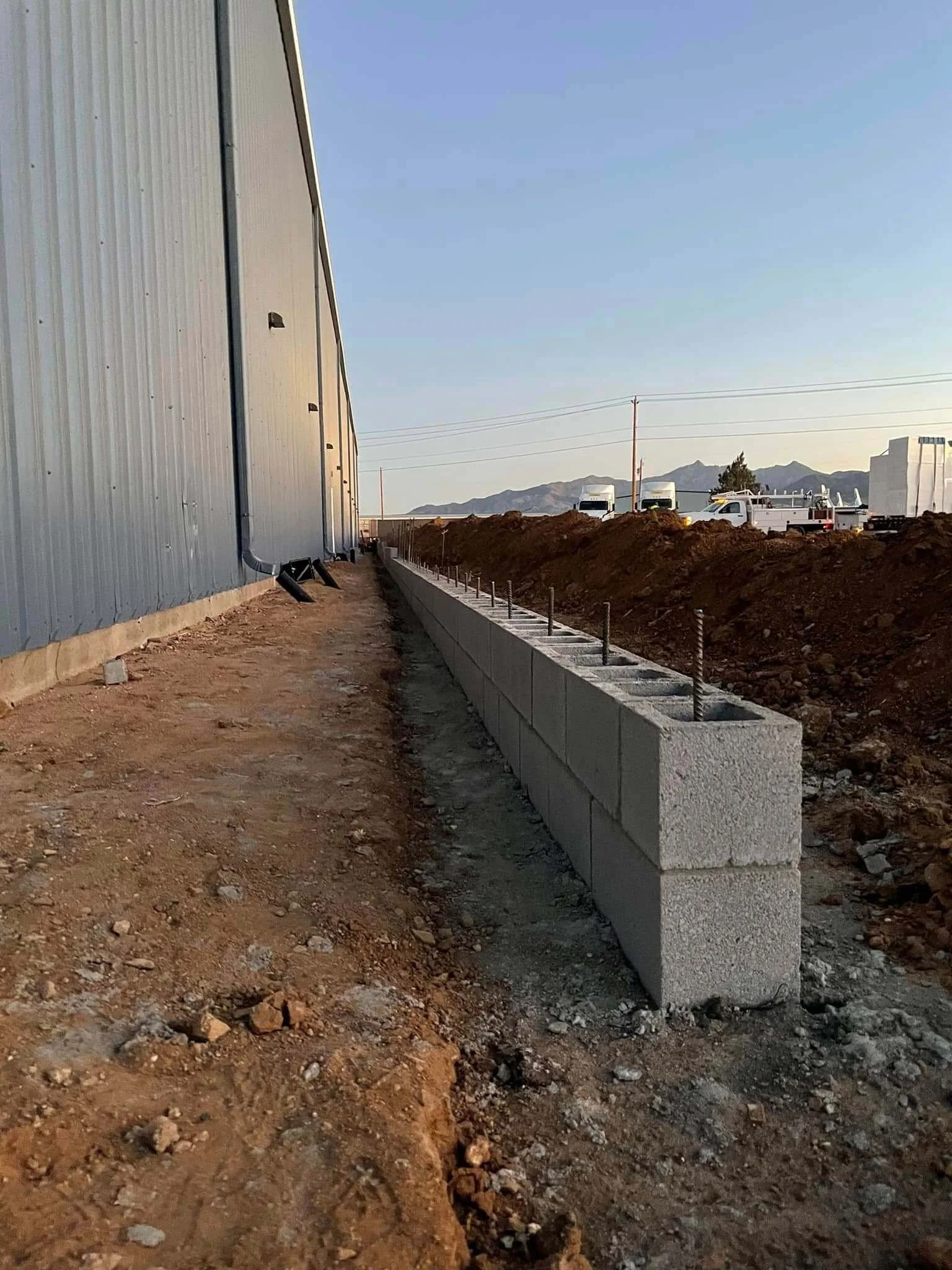 Construction site with a metal building on the left, a low concrete wall with metal rods on top, and vehicles parked in the distance with mountains in the background.