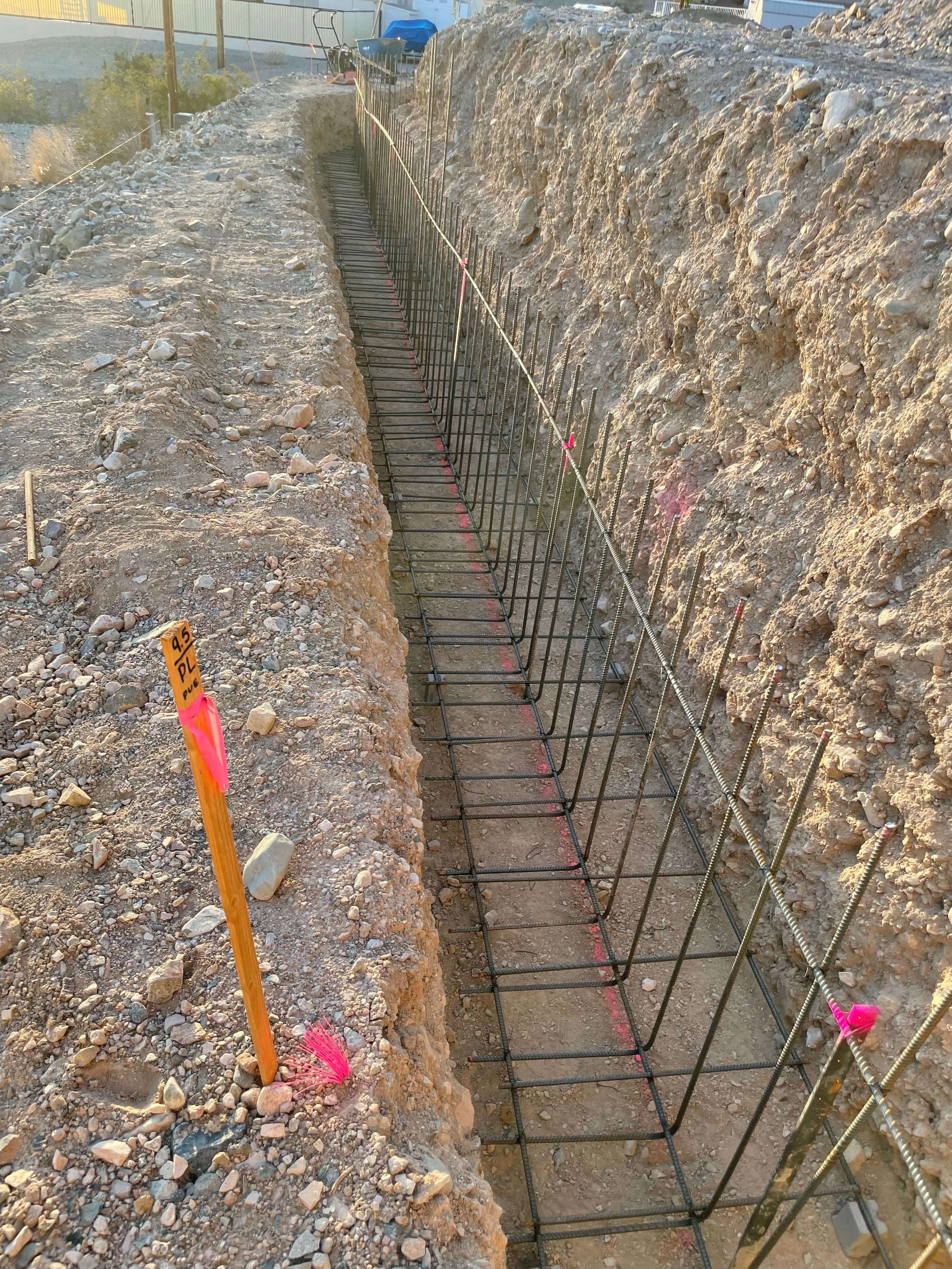 Construction site showing a trench with rebar reinforcement, gravel and rocks on the ground, and orange marking stick with pink flag attached.