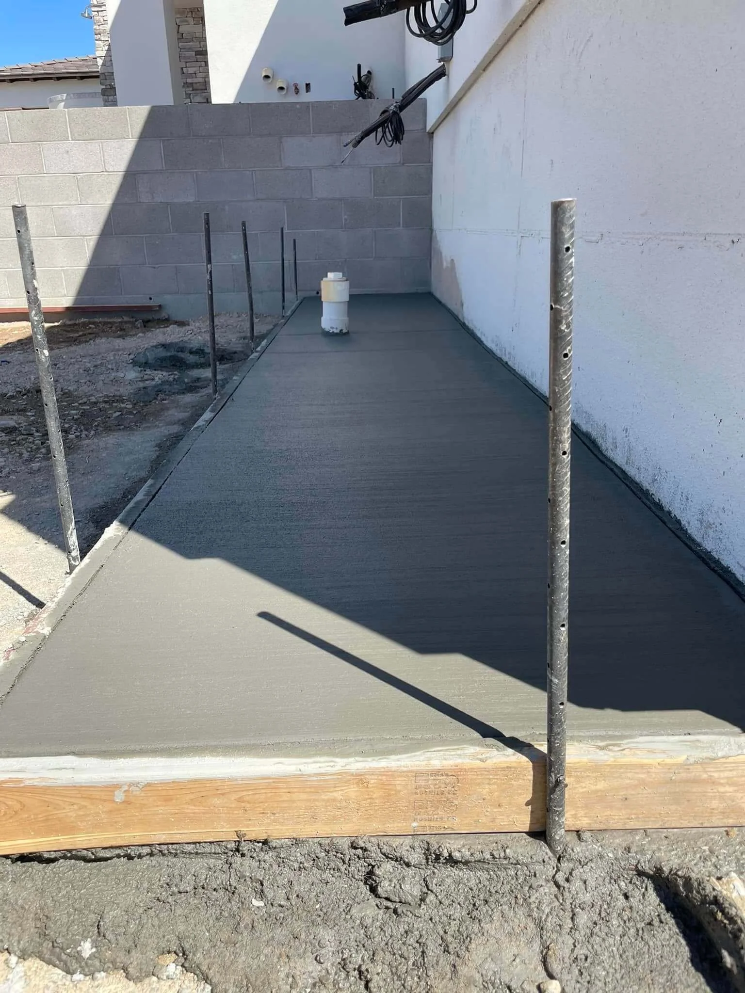 Newly poured concrete sidewalk with metal stakes for formwork, adjacent to a wooden border, in front of a building with a brick wall and white stucco exterior. Construction materials and tools are visible in the background.