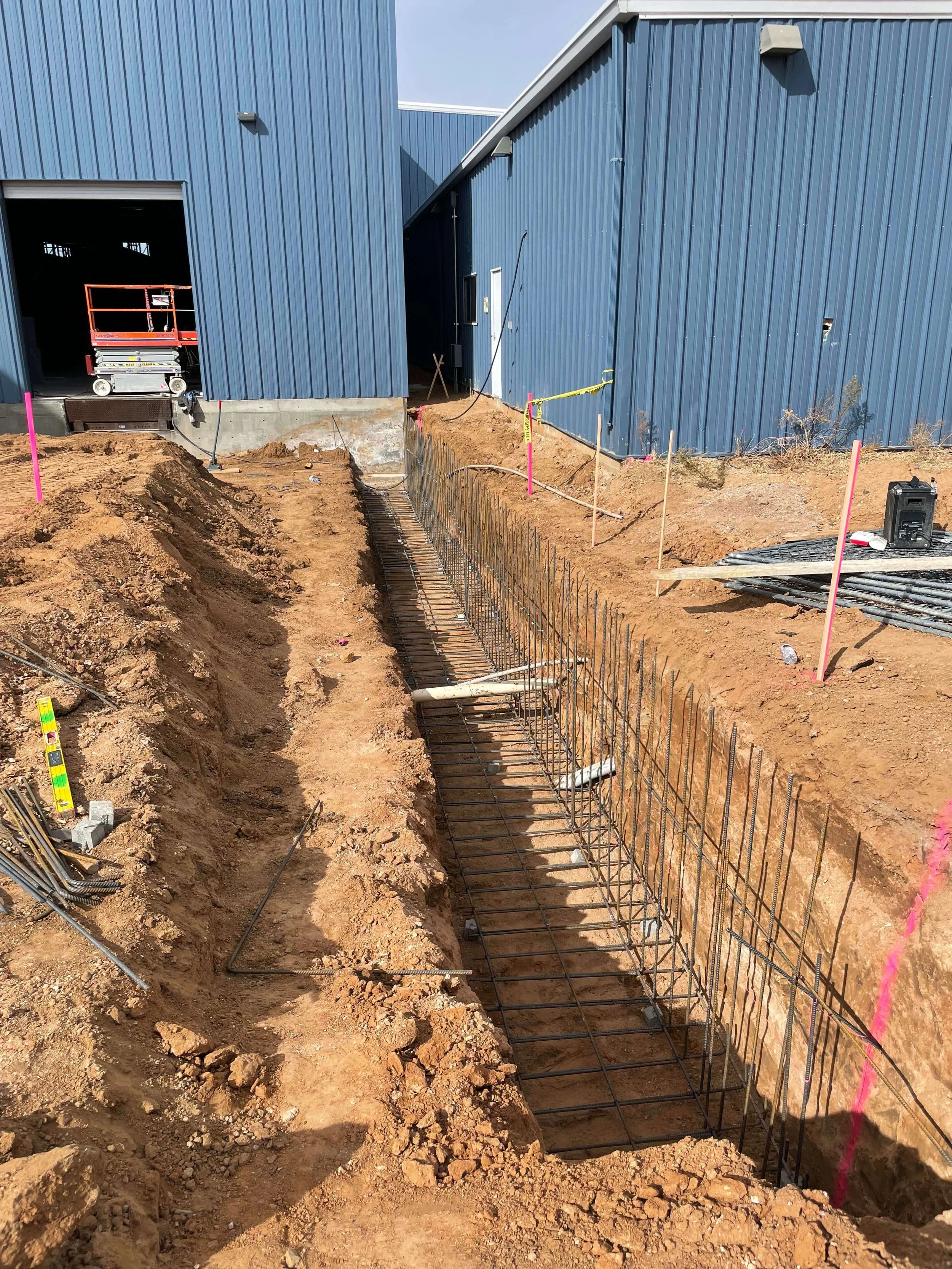 Construction site with dug trenches, metal rebar framework for concrete, and a blue building in the background.