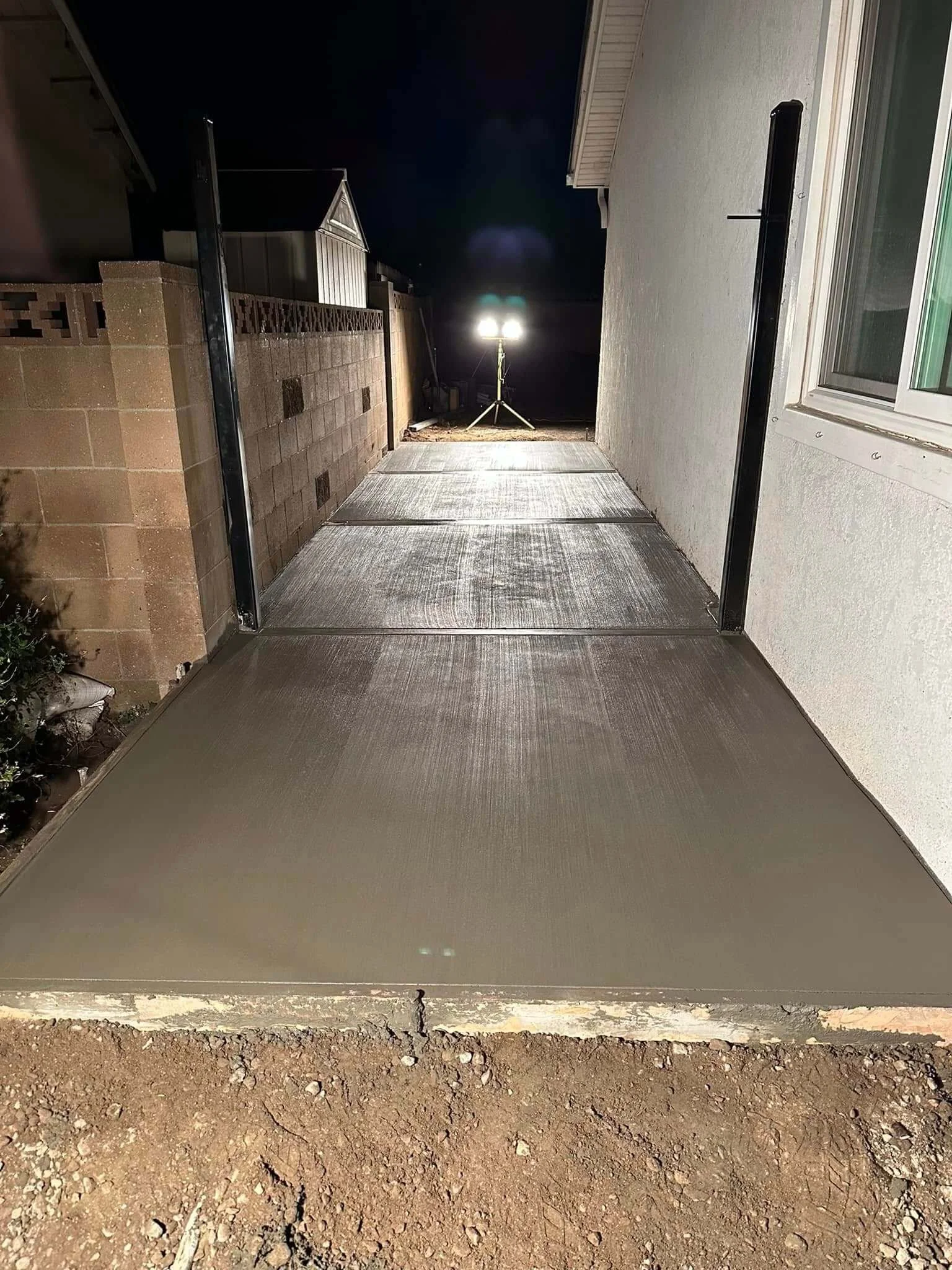 Photo of fresh concrete slab poured on ground between a house wall on the right and a brick wall on the left, with a dirt surface in the foreground and a night sky in the background.