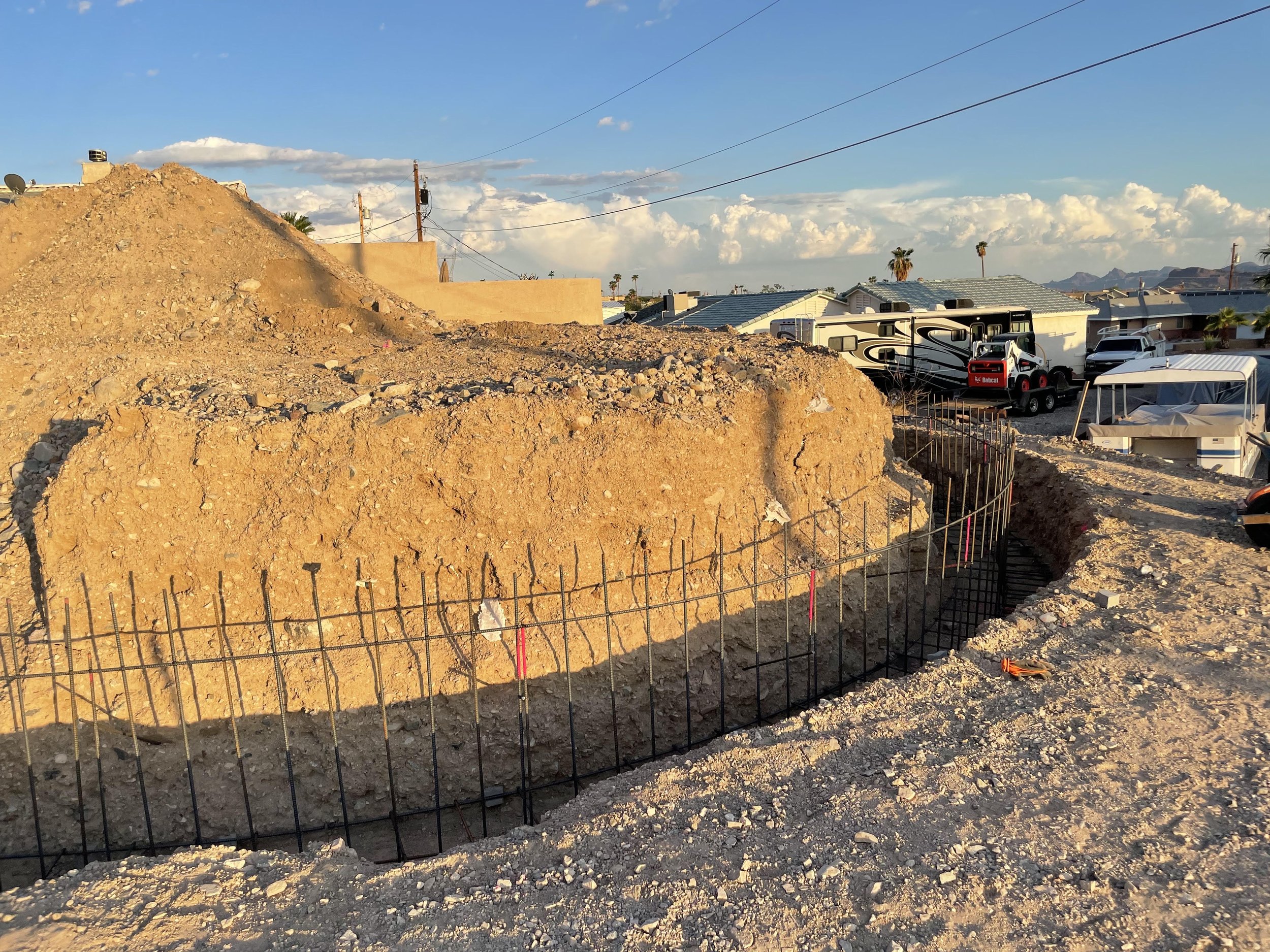 Construction site with dirt mound, fencing, and RV parked in a residential area under a blue sky with scattered clouds and mountains in the background.