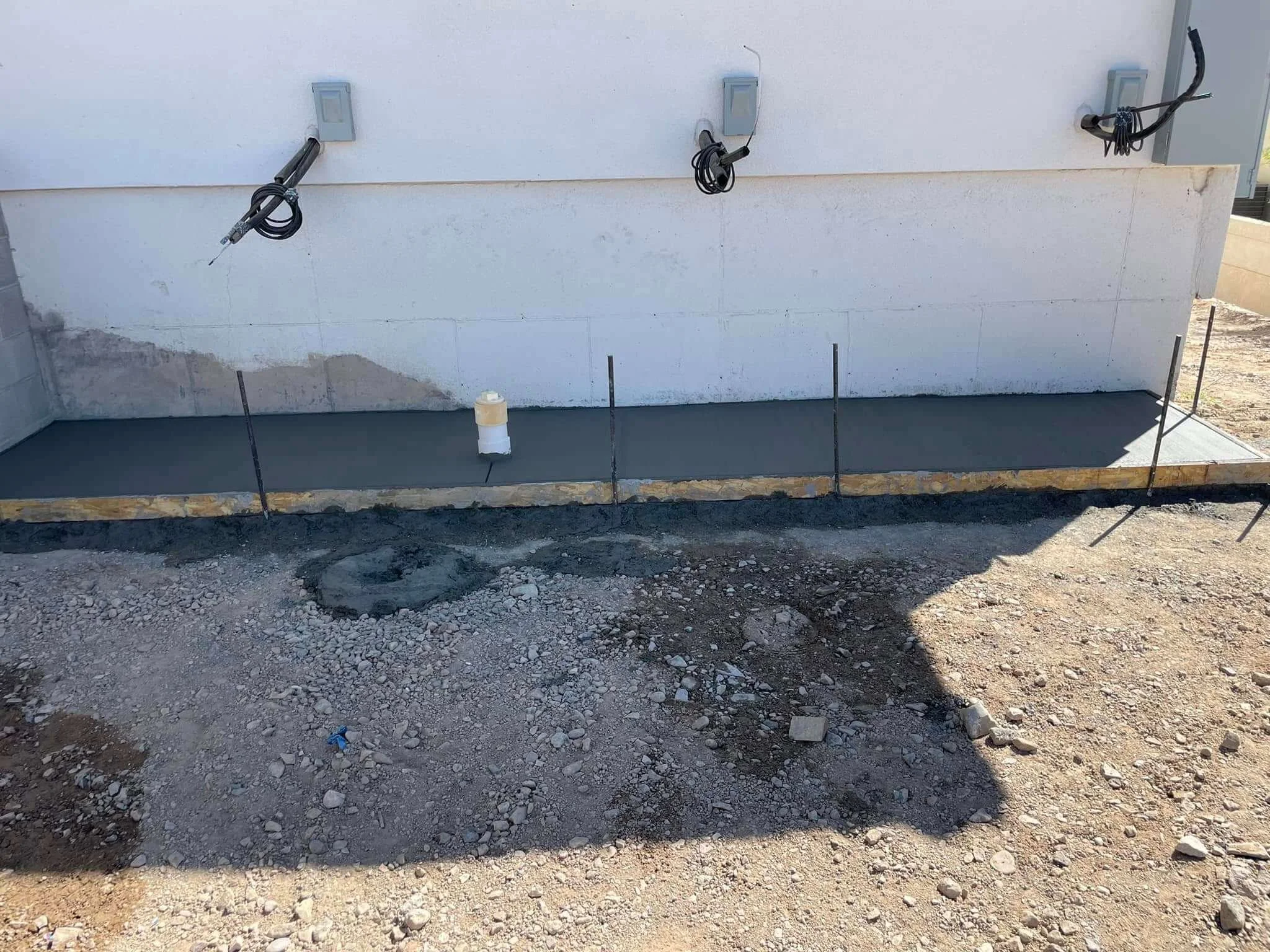 Construction site with concrete foundation and exposed pipes on a white exterior wall.