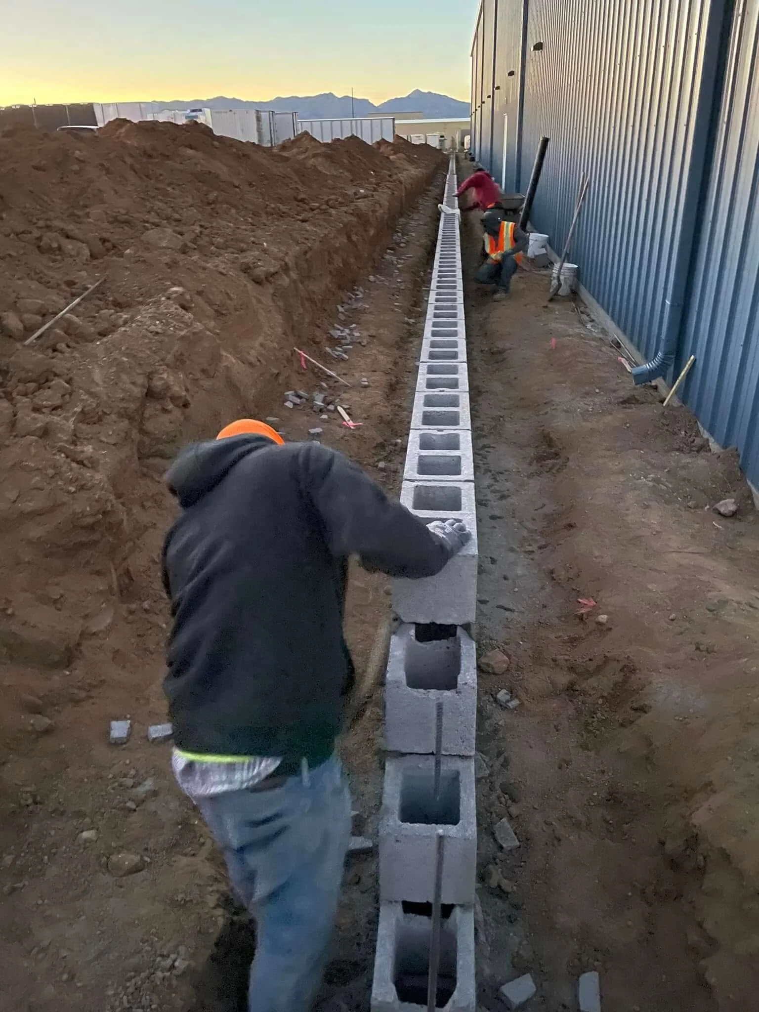 Construction workers building a concrete block wall outdoors during sunset, with dirt mounds on one side and a metal building wall on the other.