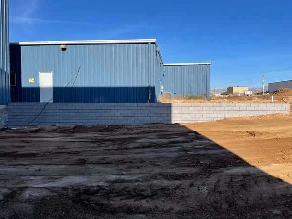 A construction site with a blue industrial building, a cinder block wall, and dirt ground under a clear blue sky.