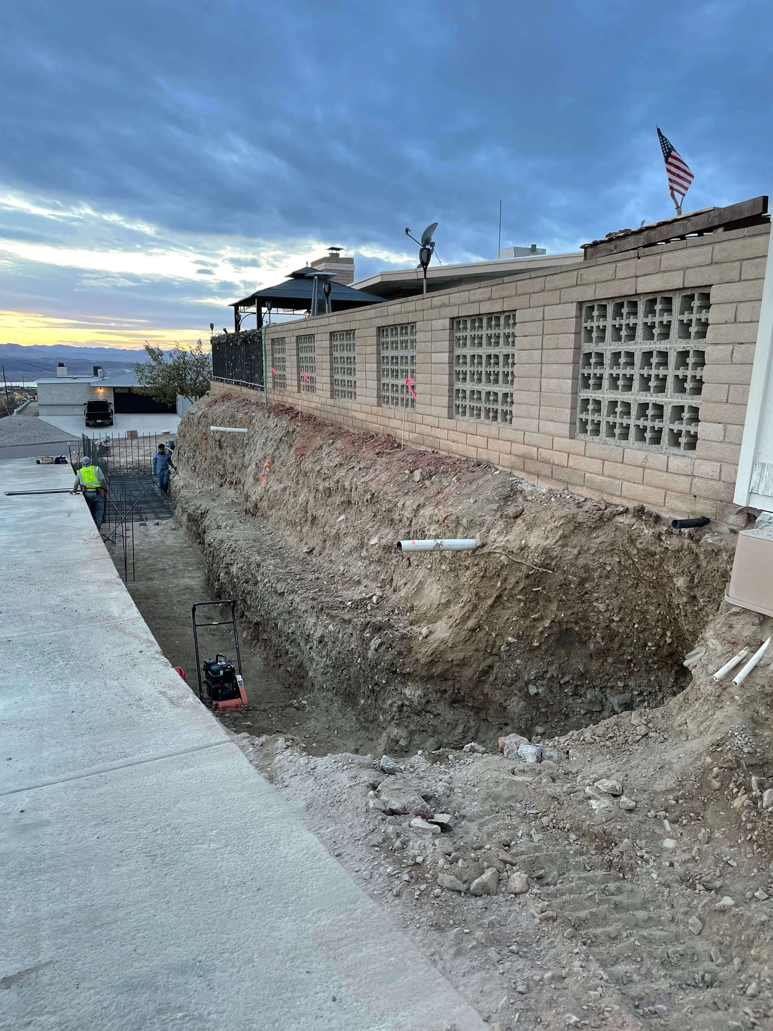 Construction workers working on a sidewalk and a retaining wall, with an excavated trench filled with dirt and rocks, in a residential area during sunset or early evening.
