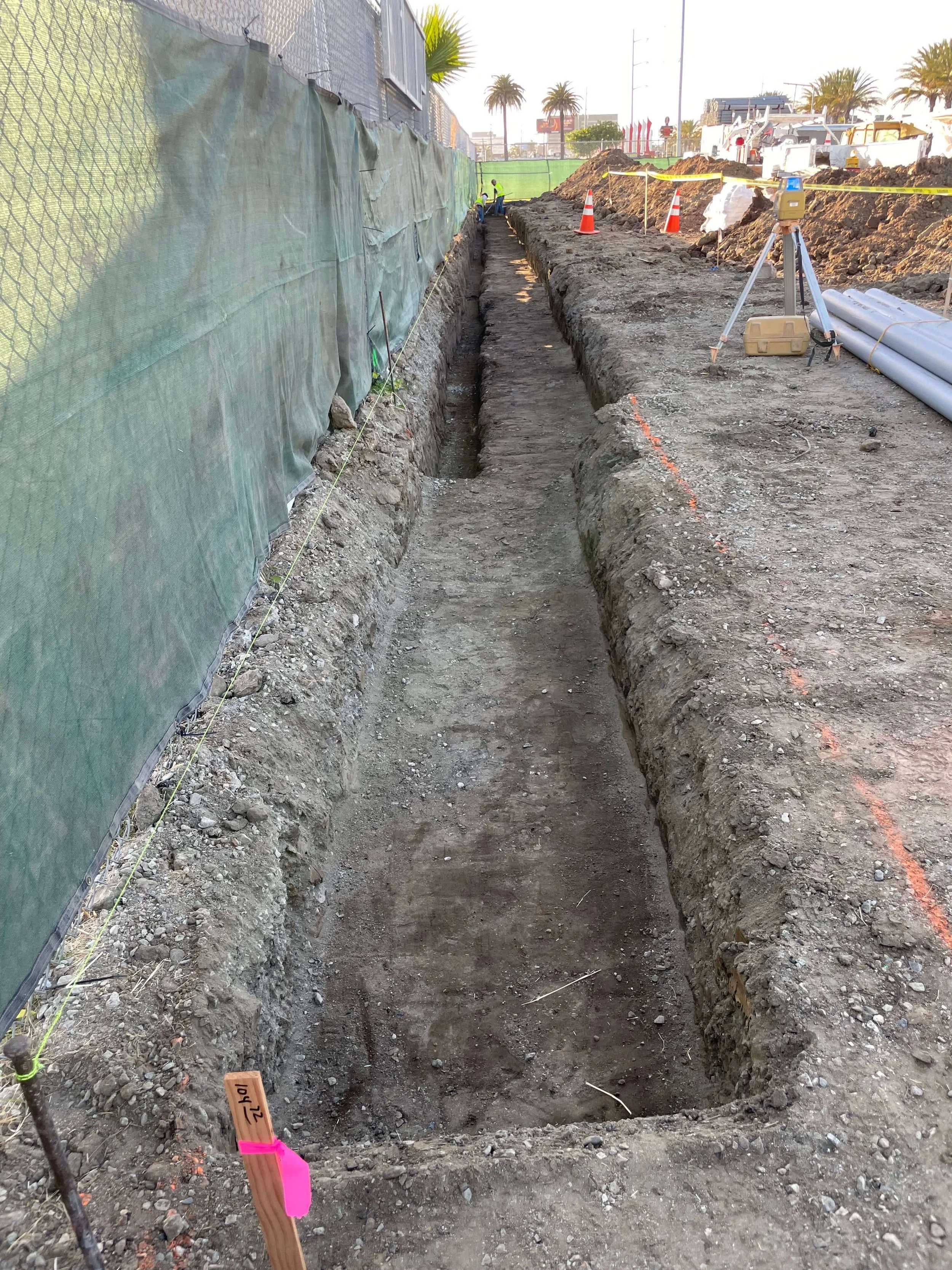 A construction site showing an excavated trench with workers and construction equipment in the background. Safety cones and a surveyor's tripod are also visible.