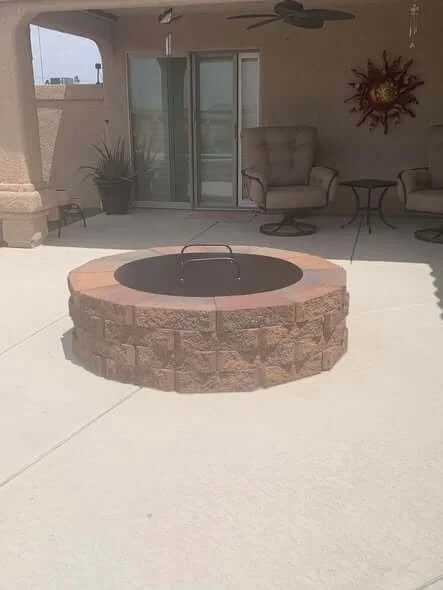 Outdoor patio with a circular fire pit made of brick and stone, accompanied by patio furniture, potted plants, and a decorative wall art piece.