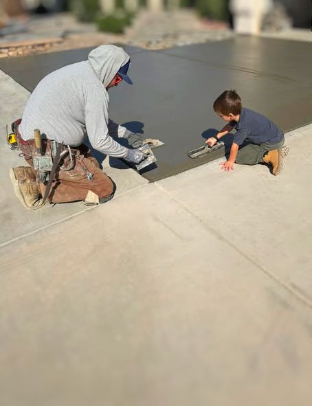 A man and a young boy installing a black roofing membrane. The man is kneeling with tools, and the boy is sitting, using a tool on the membrane.