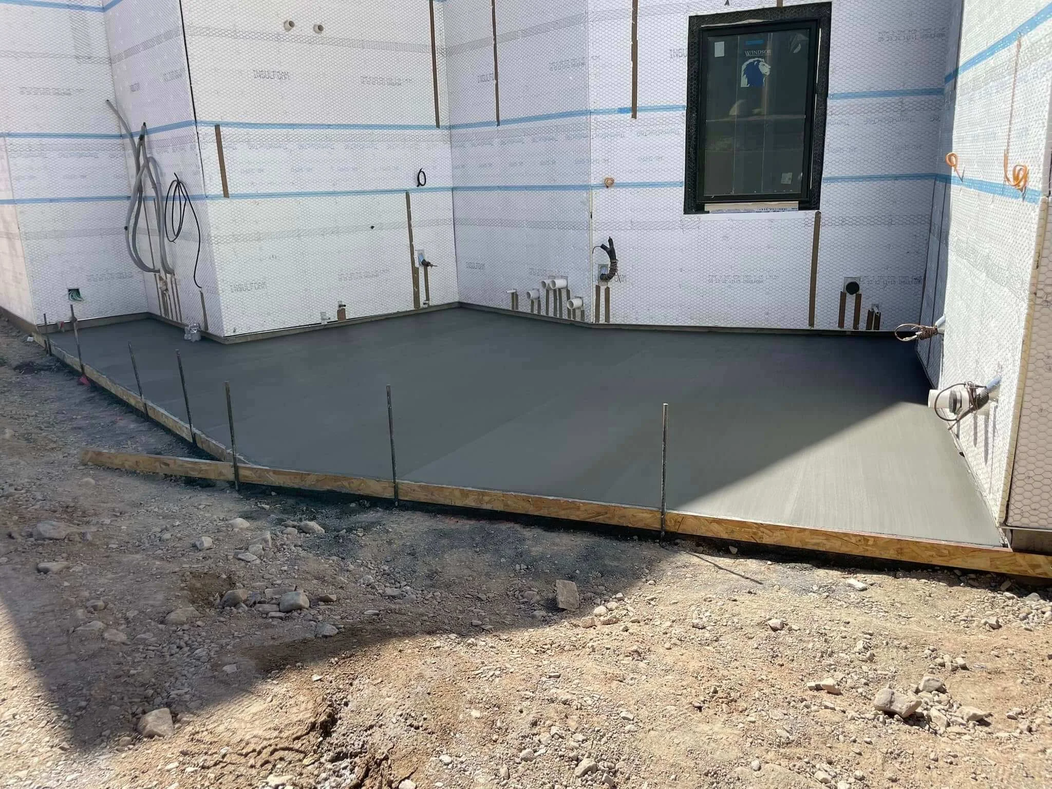 Construction site with a freshly poured concrete foundation and exposed plumbing and electrical wiring, adjacent to a partially built wall with a window.