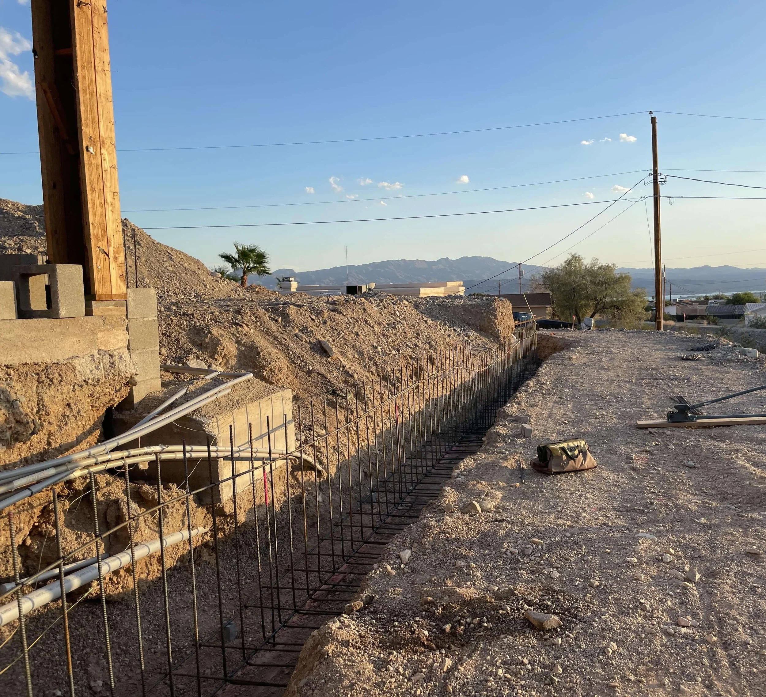 View of a construction site with a partially built wall, electrical pipes, a wooden utility pole, mountains in the background, and clear blue sky.