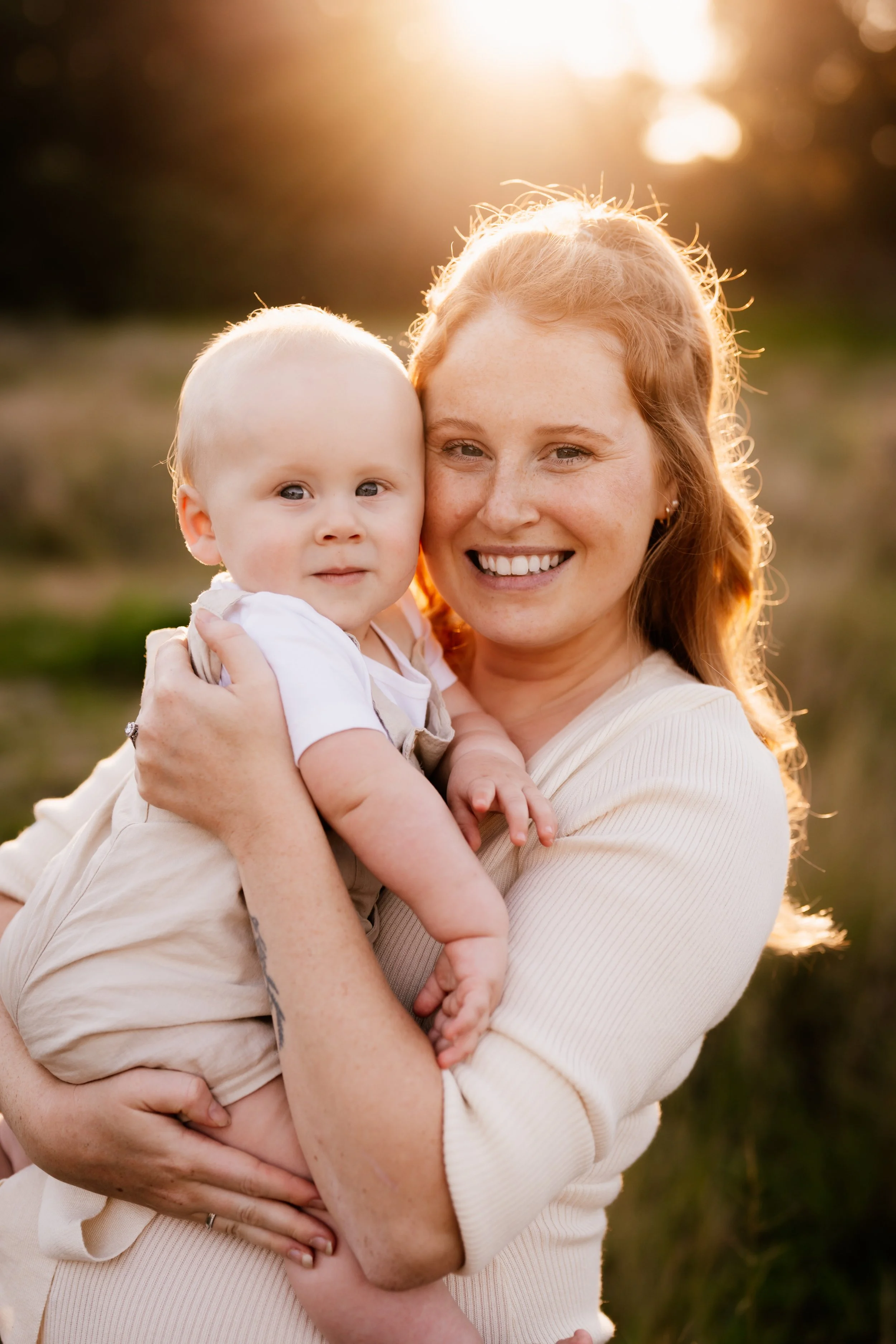 A woman with red hair and freckles is smiling and holding a young boy with blonde hair and blue eyes in her arms outdoors during sunset.