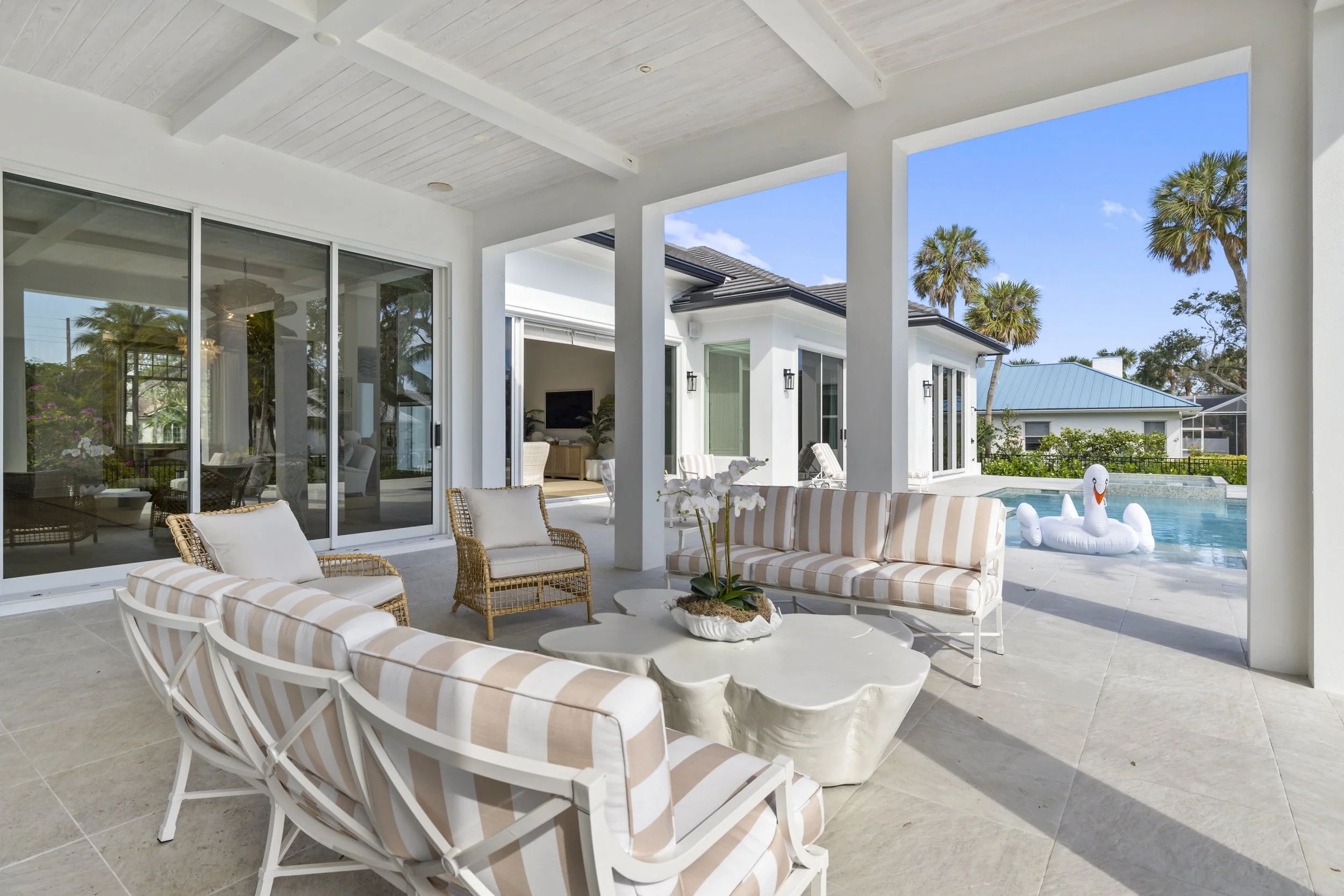 Patio area with striped outdoor furniture, potted plant, and swimming pool with inflatable swan float, surrounded by palm trees and a white house with large glass doors and windows under a blue sky.
