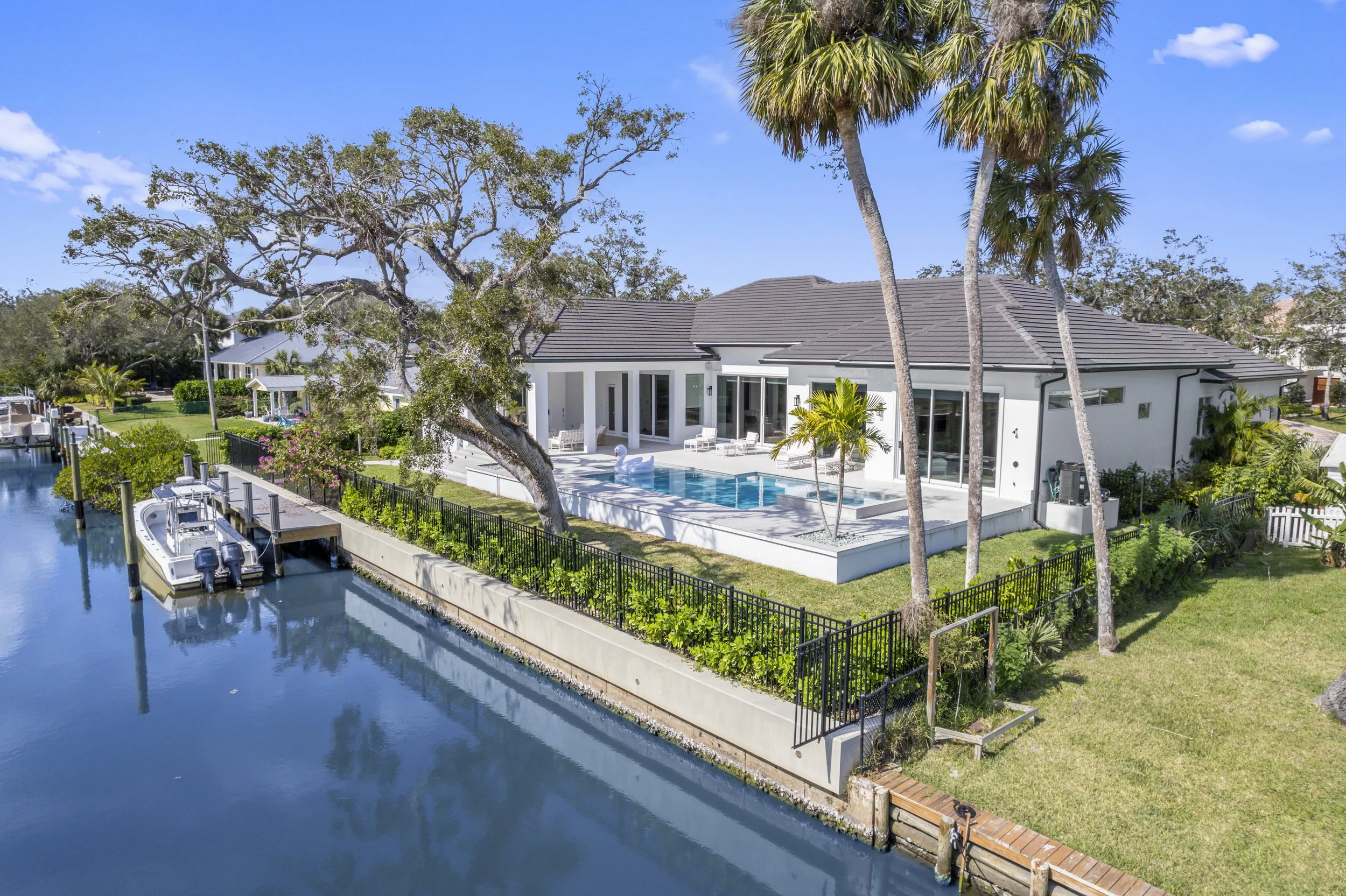 A modern house with a gray tiled roof and large windows, surrounded by lush trees including palm trees and a large oak, with a backyard swimming pool and patio furniture. There is a boat docked along a canal beside the property under a blue sky with scattered clouds.