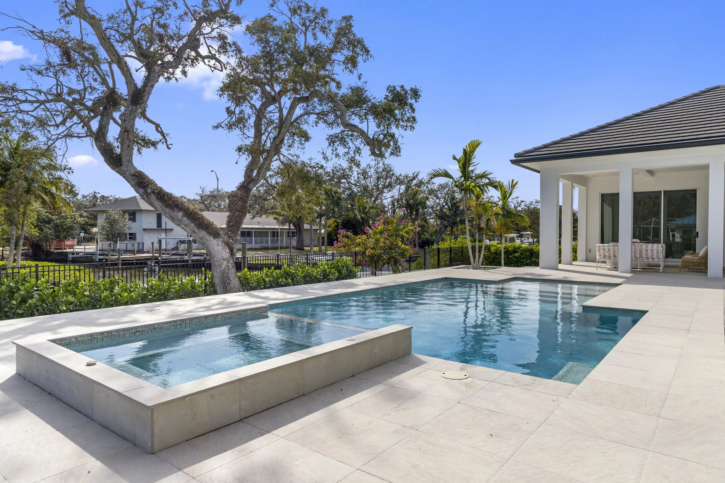 Luxury backyard with swimming pool, hot tub, patio area, and house with porch. Large tree and lush bushes in the background, sunny sky.