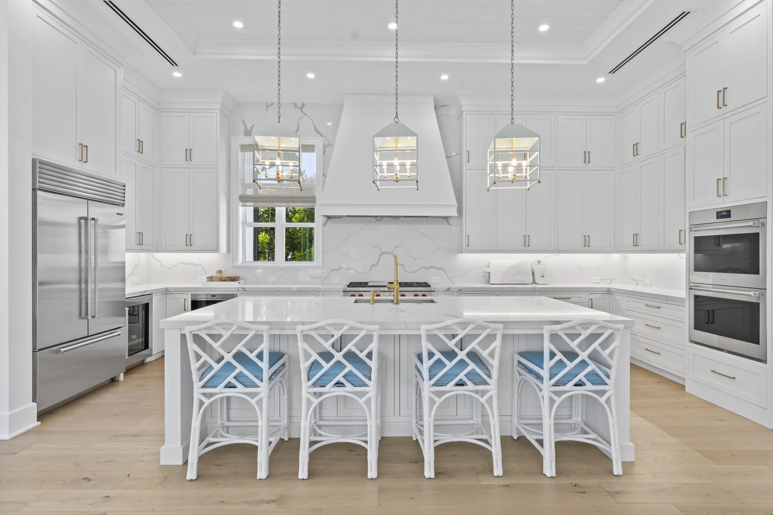 Modern white kitchen with island, pendant lights, and built-in appliances, hardwood floors, and a window showing outdoor greenery. Professional real estate photo taken by Lee at PH MEDIA of a home in vero beach florida