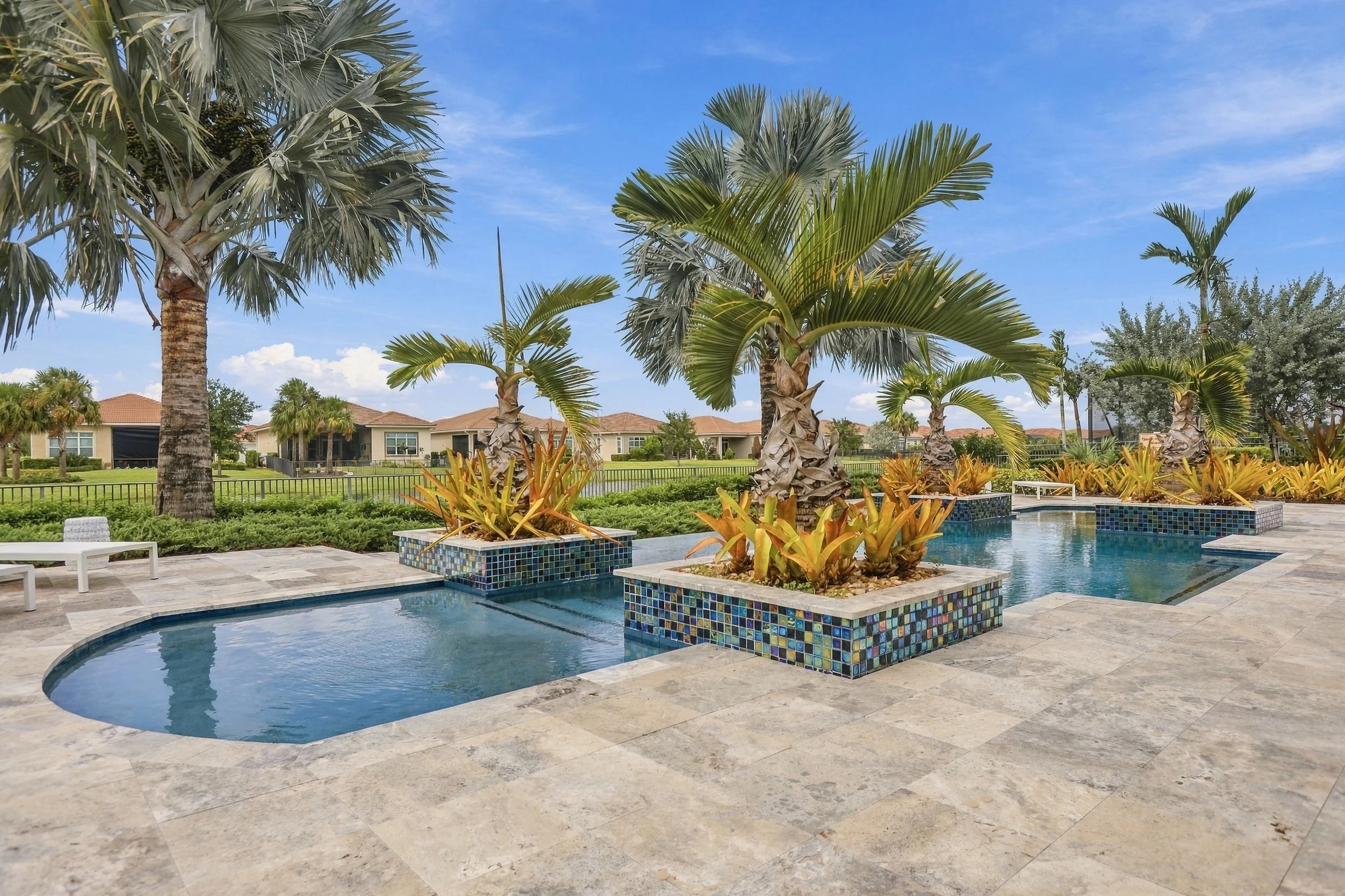 View of a luxurious swimming pool area with colorful tiled planters filled with palm trees and plants, surrounded by a patio with lounge chairs, and a backdrop of residential houses and clear blue sky.