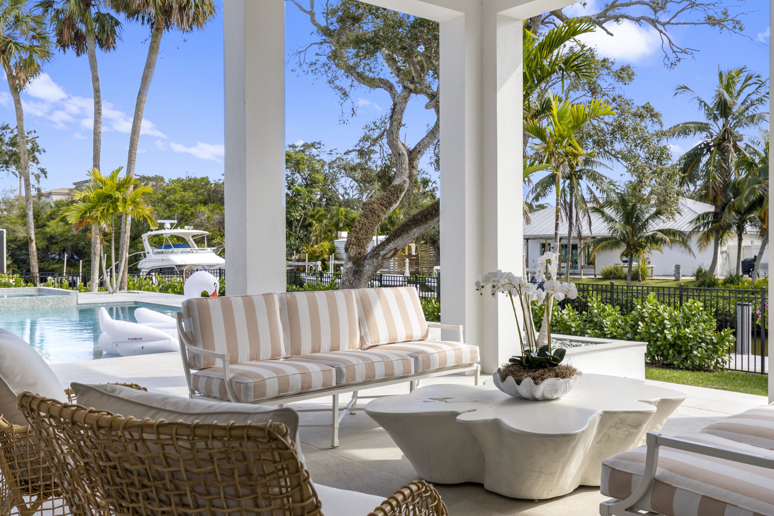 Professional real estate photo taken by Lee. view from a covered outdoor seating area with striped cushions, overlooking a backyard with a swimming pool, a docked boat, lush trees, and a white house with a metal roof in the background on a sunny day.